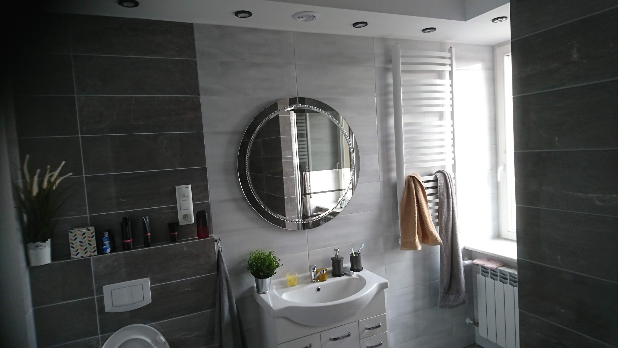 Nowoczesna łazienka z szarymi płytkami, okrągłym lustrem z ozdobną ramą, białą umywalką z szafką i grzejnikiem drabinkowym z ręcznikami.
