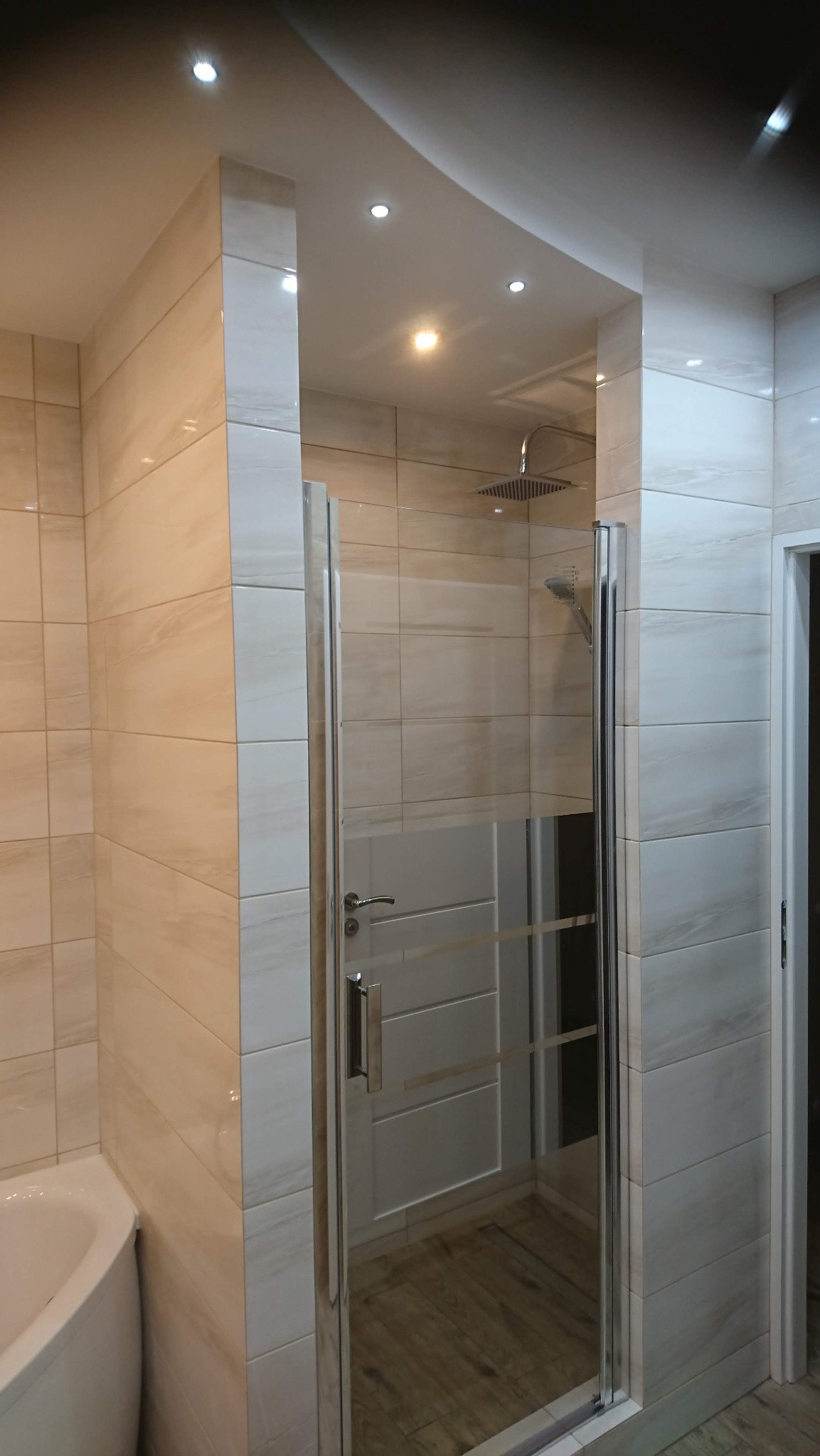 Nowoczesna kabina prysznicowa z przesuwnymi drzwiami, wykończona beżowymi płytkami imitującymi marmur, z widoczną deszczownicą i oświetleniem punktowym.