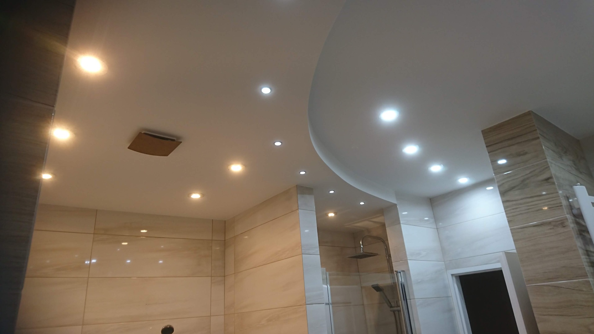 Wykończona łazienka z podwieszanym sufitem z oświetleniem punktowym, beżowymi płytkami imitującymi marmur na ścianach i kabiną prysznicową typu walk-in.