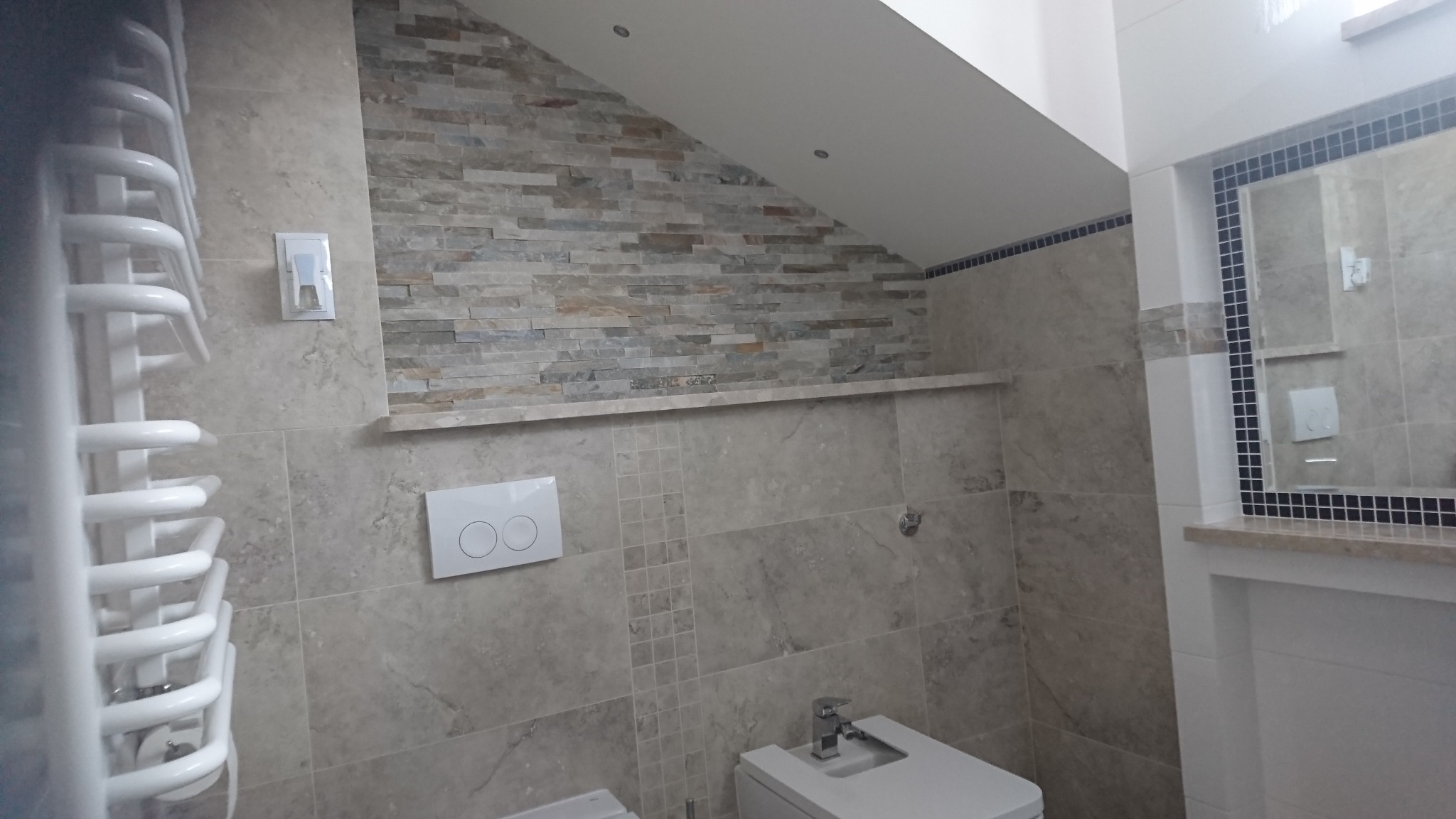 Wykończona łazienka w jasnych odcieniach beżu i szarości, z kamienną mozaiką na skośnej ścianie, grzejnikiem drabinkowym po lewej i lustrem w czarnej ramce po prawej.