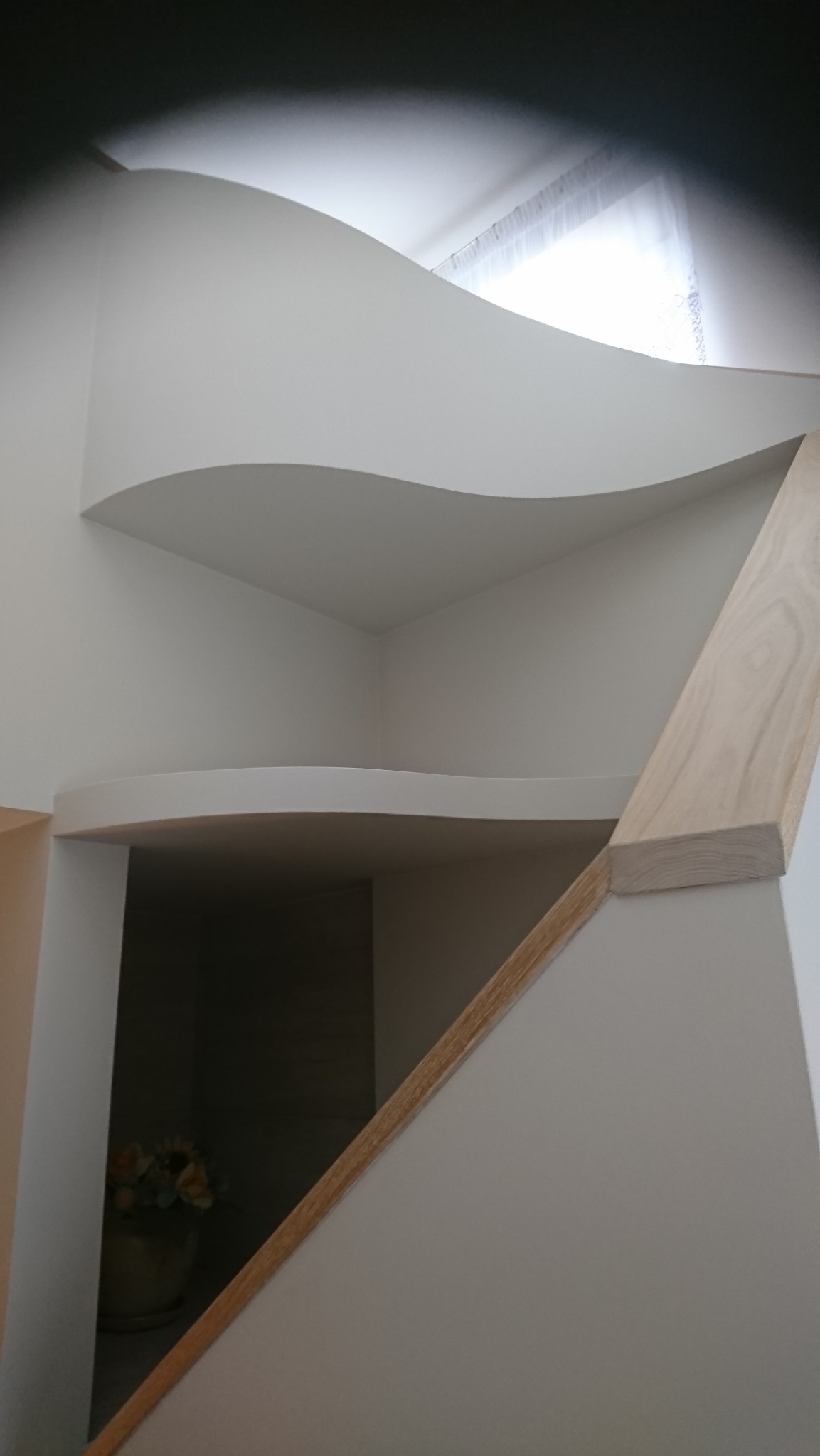 Nietypowa, falista zabudowa z płyt karton-gips w jasnym wnętrzu, z widoczną drewnianą listwą i kwiatami w tle.