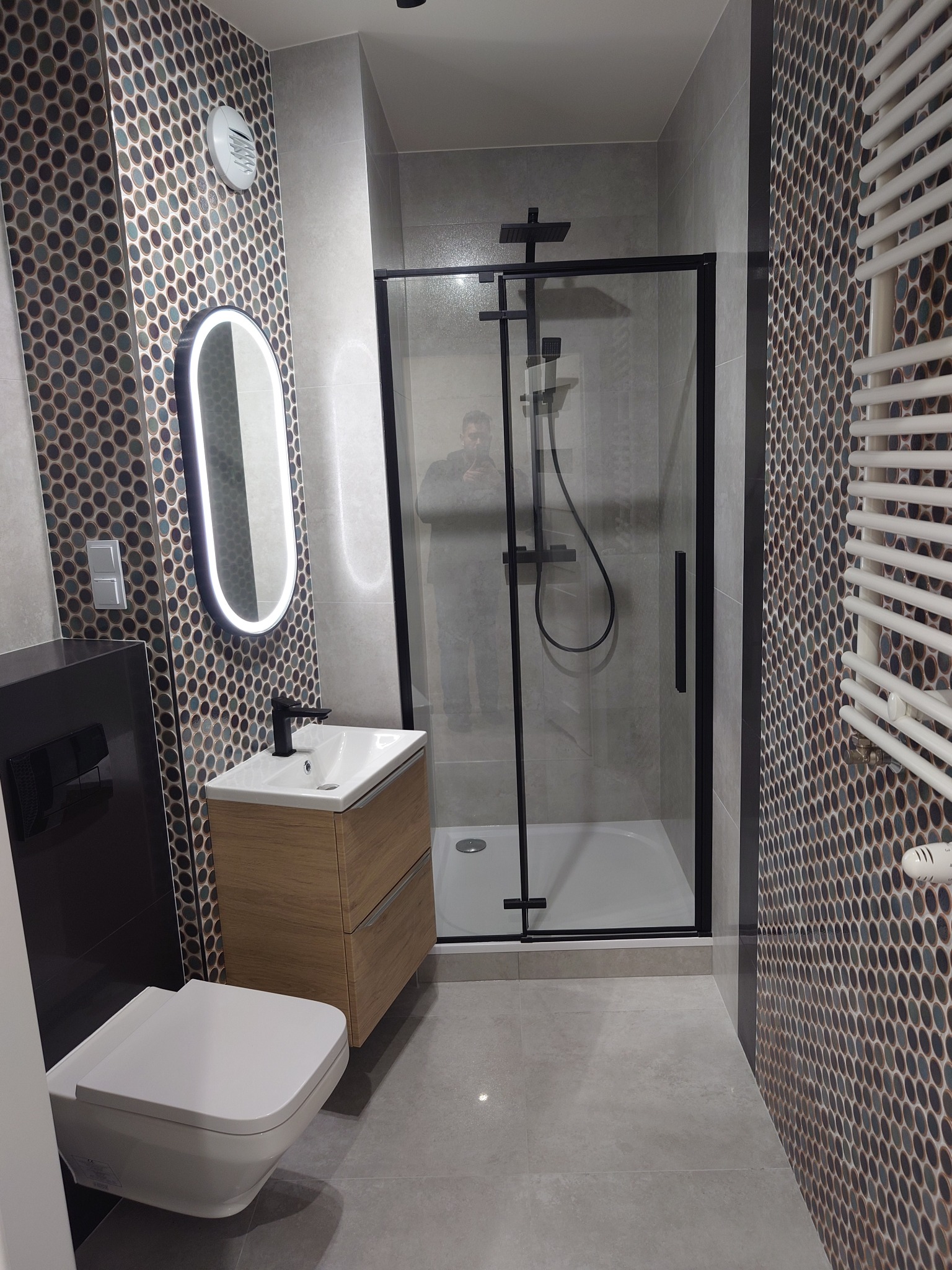 Nowoczesna łazienka z kabiną prysznicową w czarnej ramie, umywalką z szafką w kolorze drewna, podświetlanym lustrem owalnym i mozaiką na ścianach.