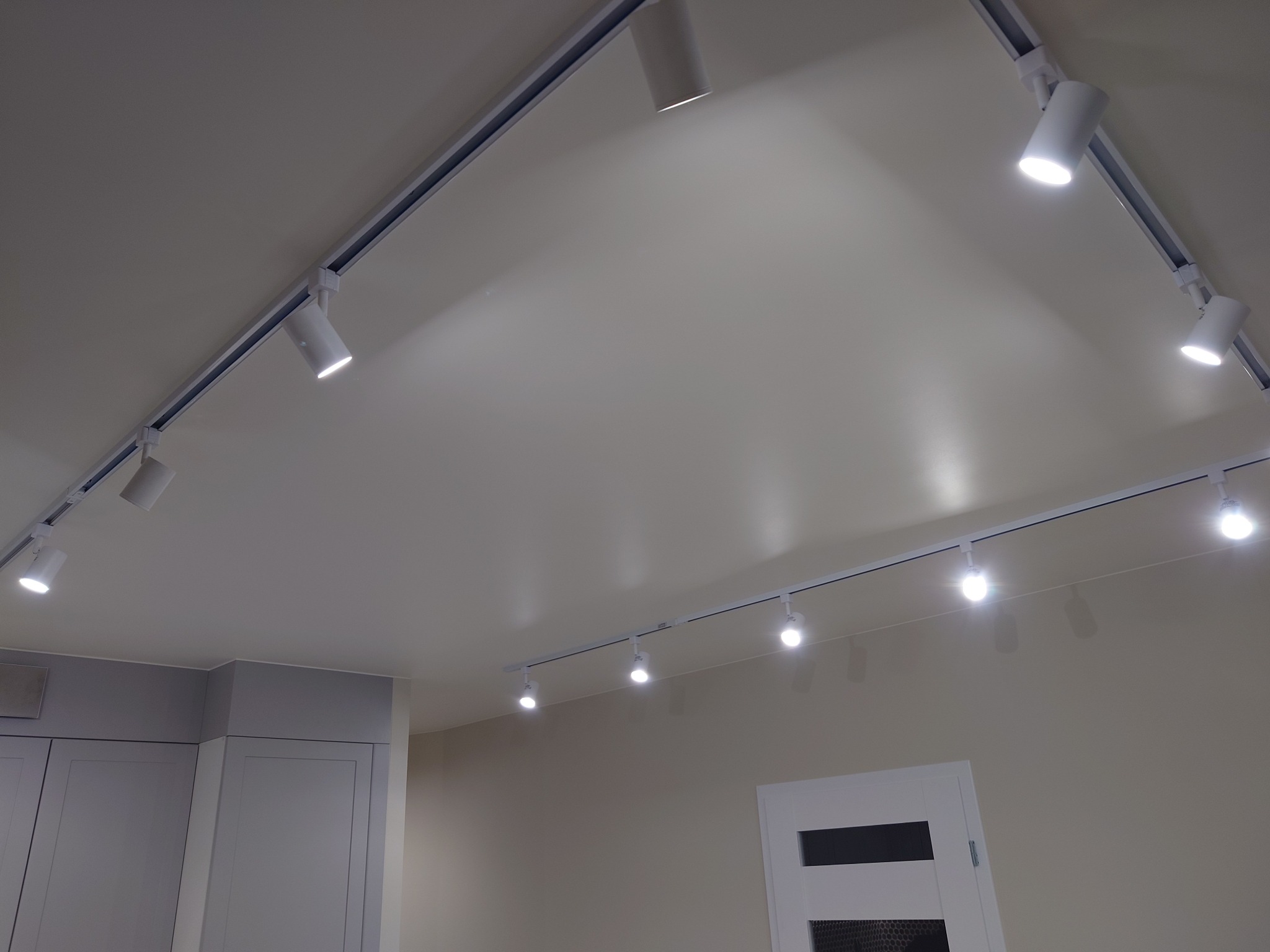 Białe reflektory szynowe zamontowane na suficie w jasnym pomieszczeniu, widoczne fragmenty zabudowy meblowej i drzwi.