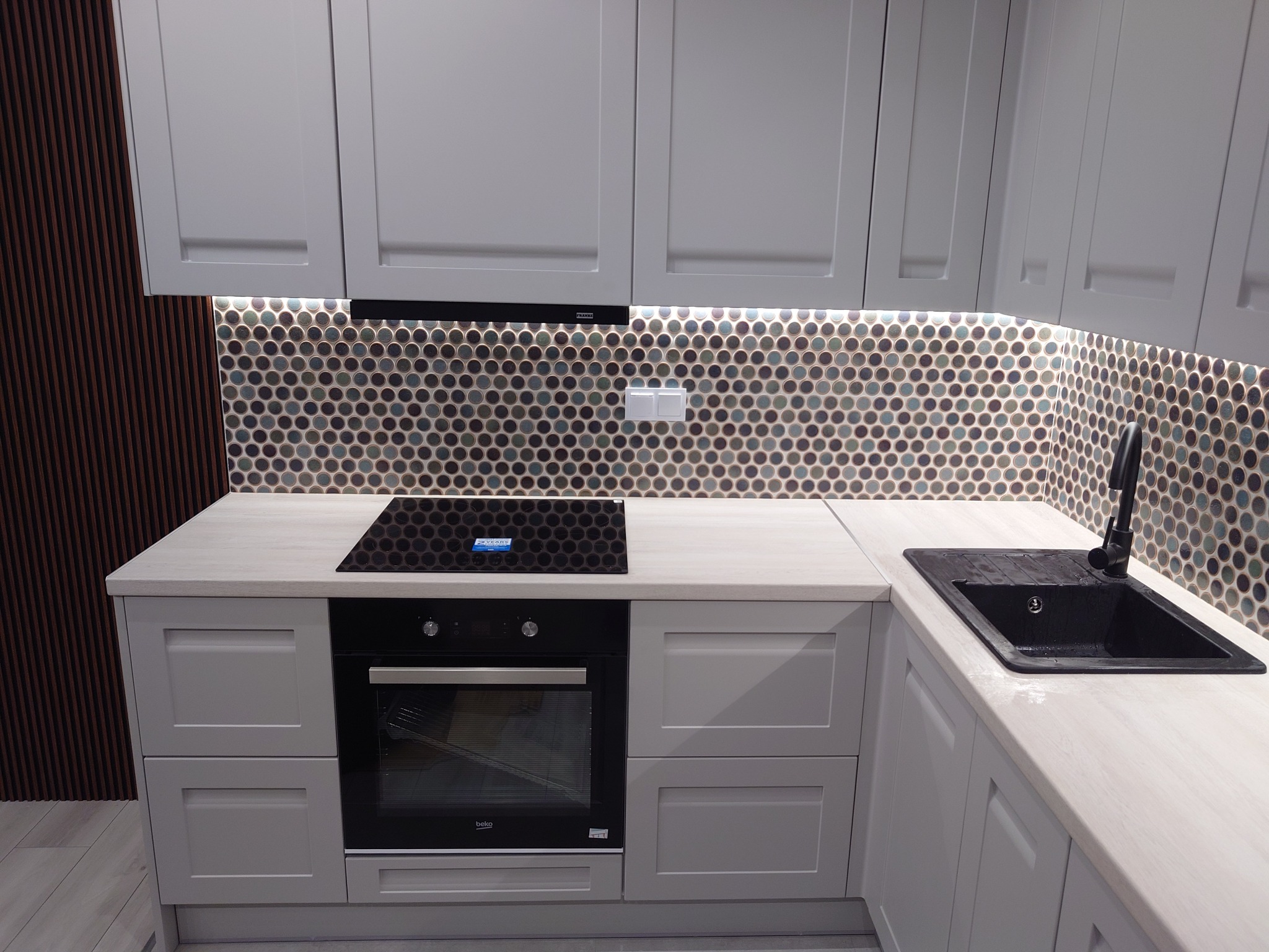 Nowoczesna, szara kuchnia z okrągłą mozaiką na ścianie, czarnym zlewem i płytą indukcyjną, oświetleniem LED pod szafkami i drewnianą okładziną ścienną