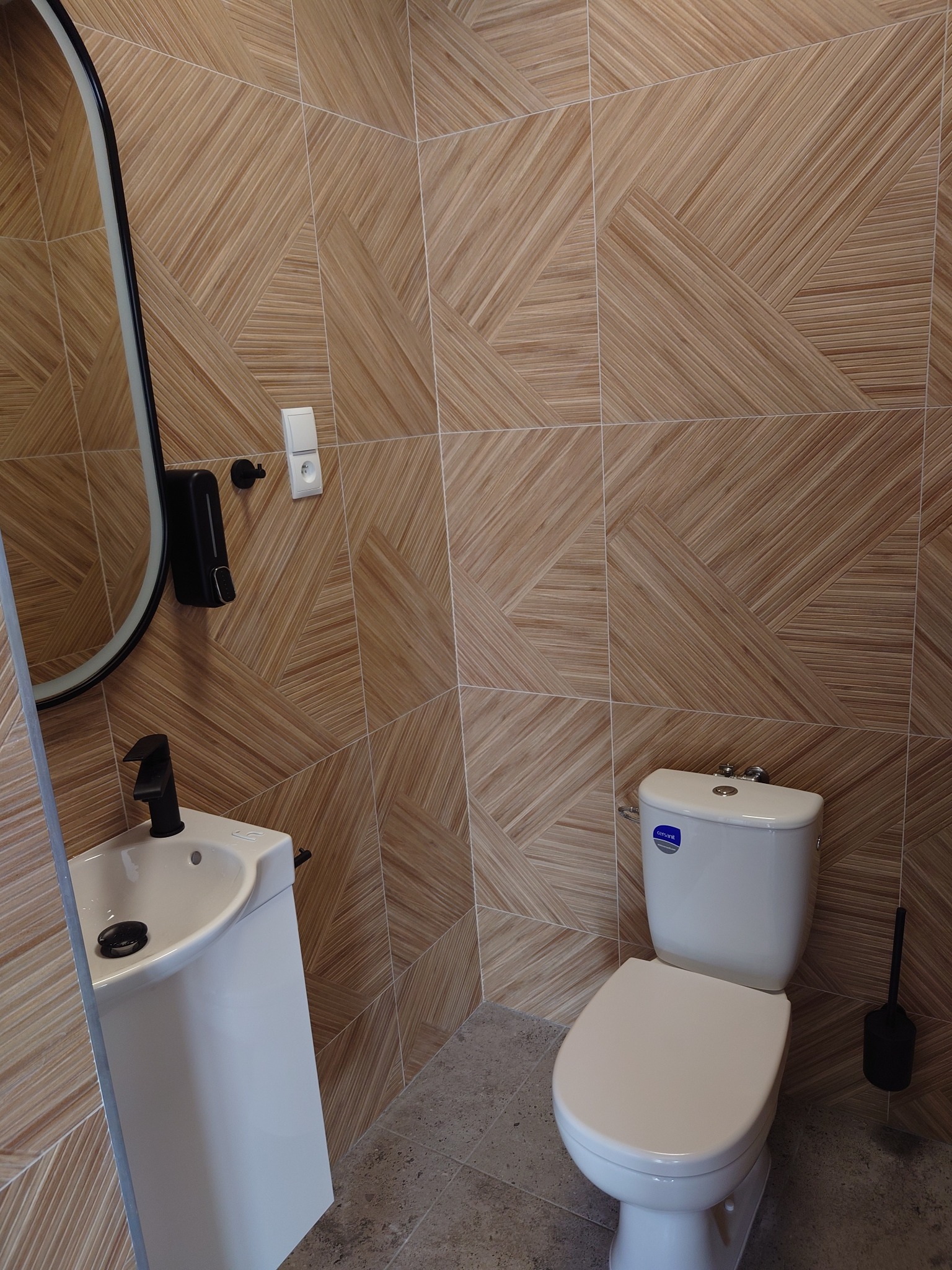 Nowoczesna łazienka z geometrycznymi, drewnopodobnymi płytkami na ścianach, białą toaletą i umywalką z czarną armaturą oraz owalnym lustrem.