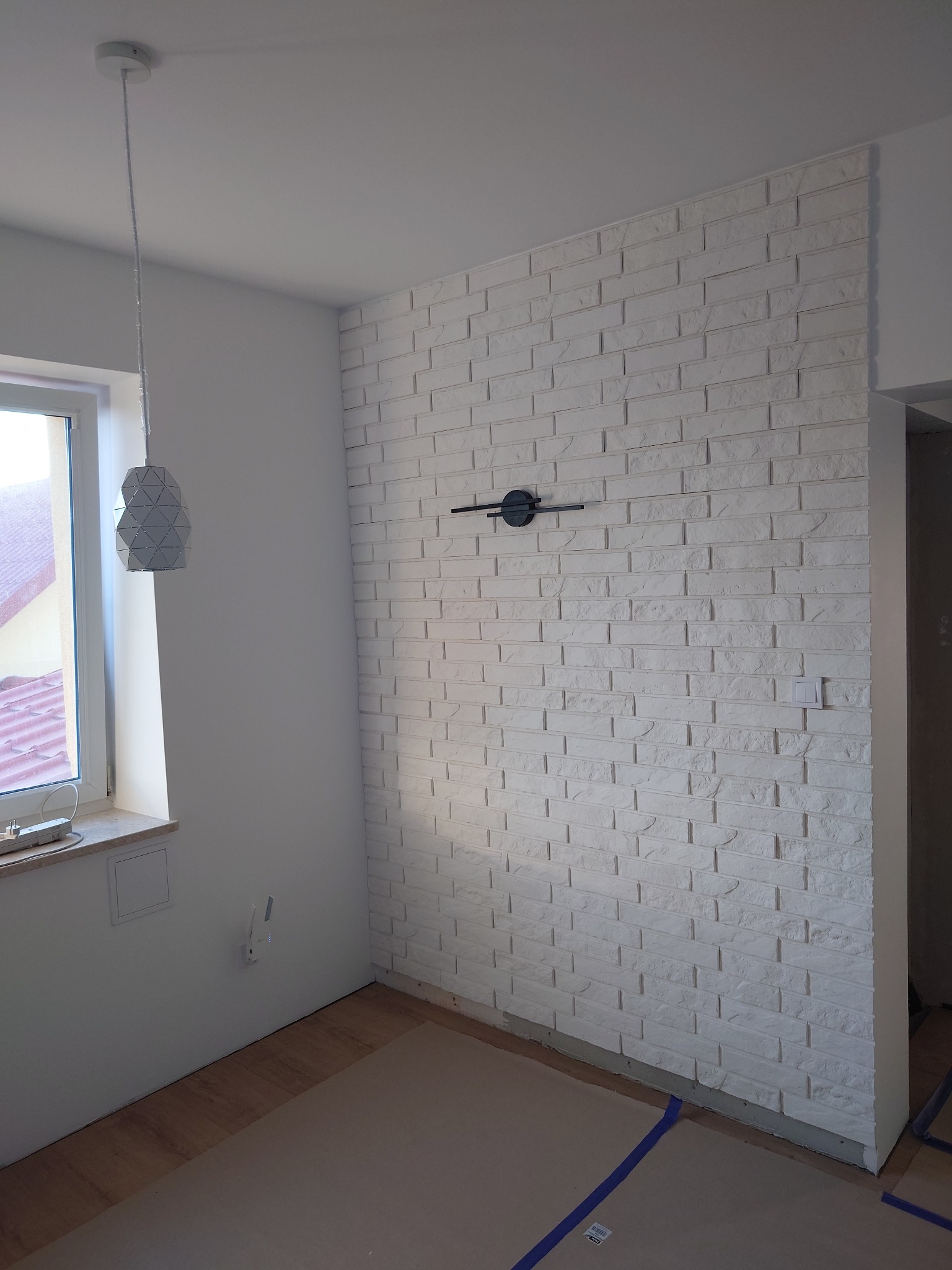 Ściana wykończona białą cegłą dekoracyjną z zamontowanym czarnym zegarem, widoczna lampa wisząca i okno, podłoga zabezpieczona kartonem i taśmą malarską.