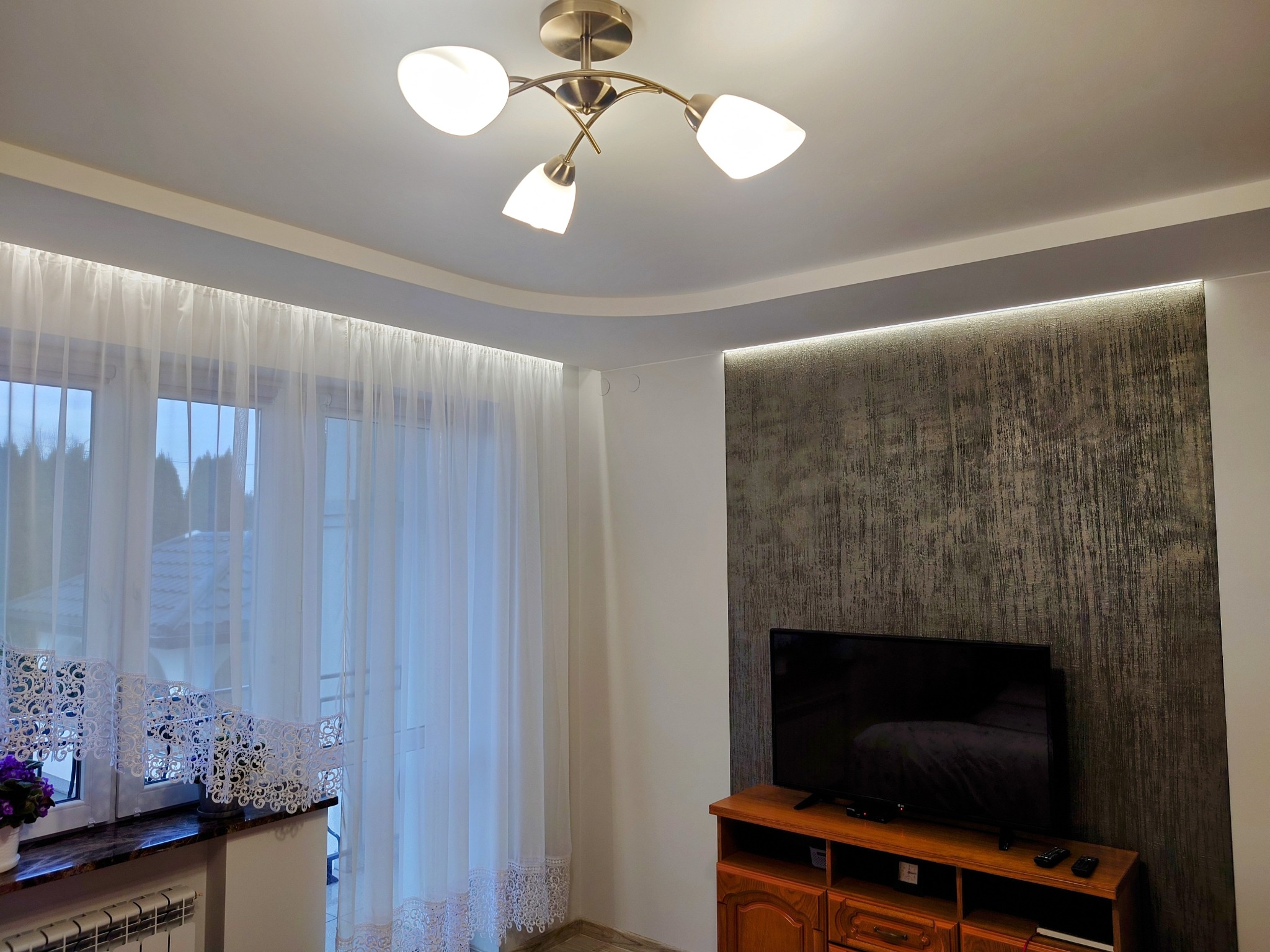 Jasny salon z dekoracyjną ścianą o fakturze betonu, oświetleniem LED podsufitowym i lampą sufitową w kolorze mosiądzu oraz telewizorem na drewnianej komodzie.