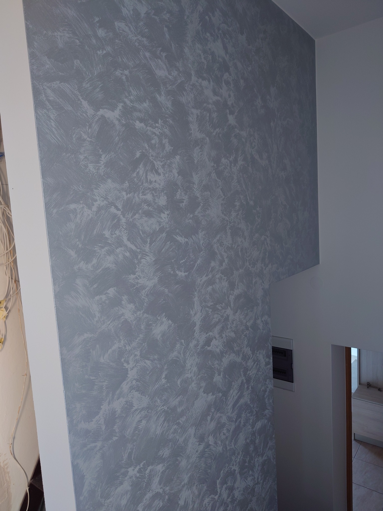 Ściana wewnątrz budynku pokryta dekoracyjną szarą farbą z efektem pociągnięć pędzla, widoczne przewody elektryczne w rogu pomieszczenia.