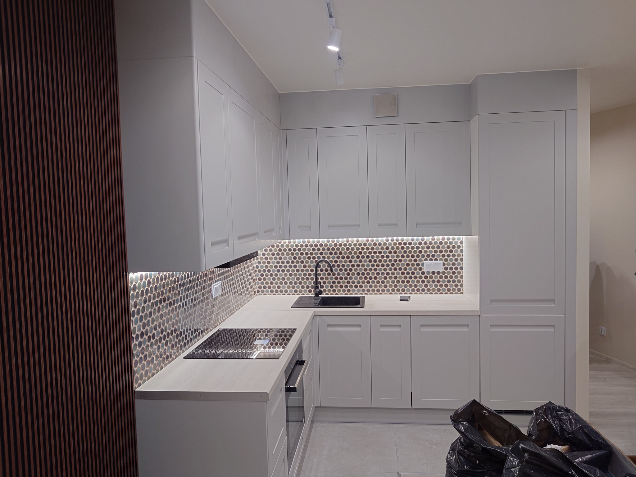 Jasnoszara kuchnia z mozaiką na ścianie, czarnym zlewem i płytą indukcyjną, widoczne listwy dekoracyjne na ścianie i worki na śmieci na podłodze.