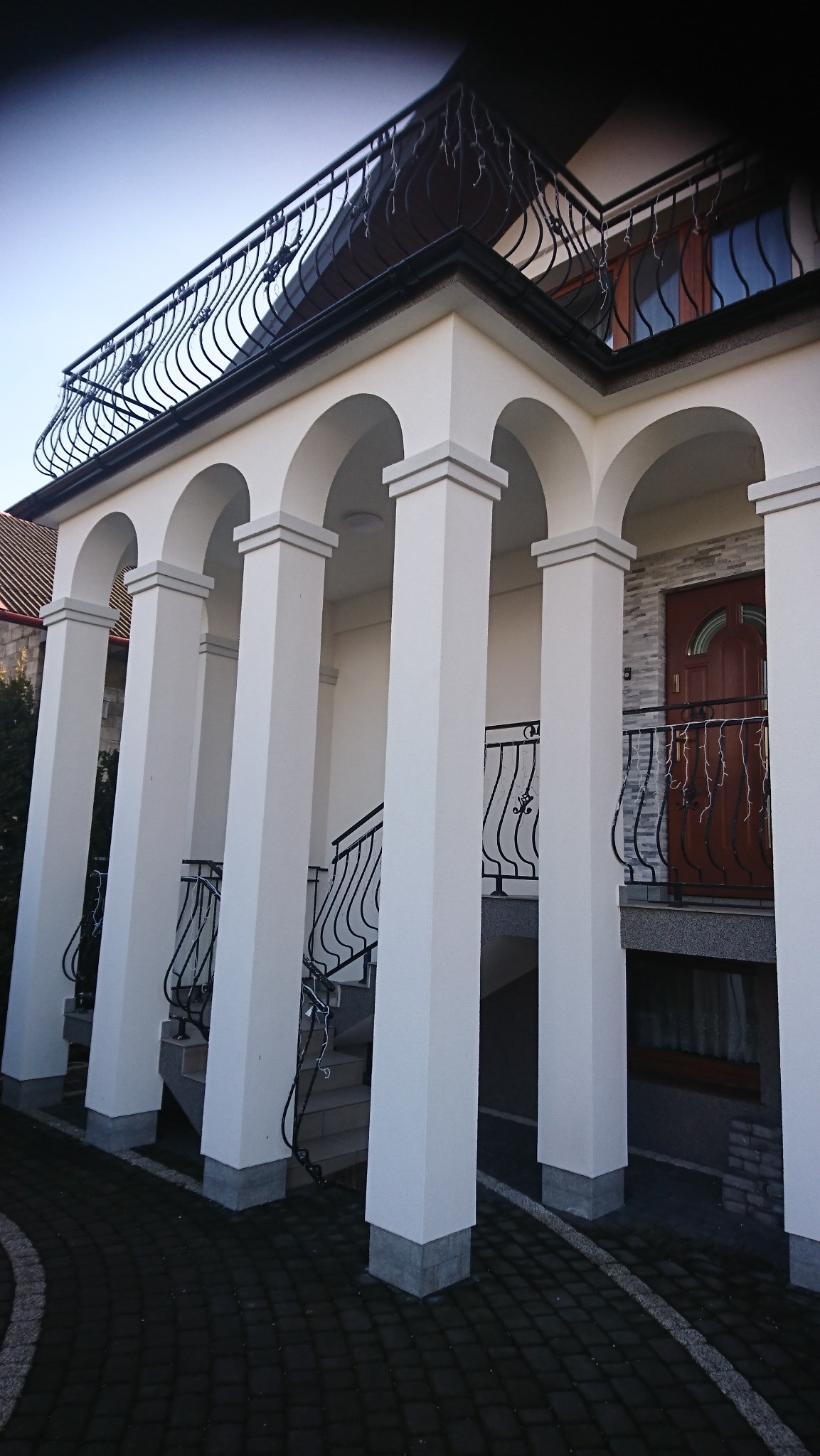 Biała kolumnada z łukami i ozdobną, kutą balustradą balkonu, wykończenie elewacji domu z drewnianymi drzwiami wejściowymi i kamienną okładziną.