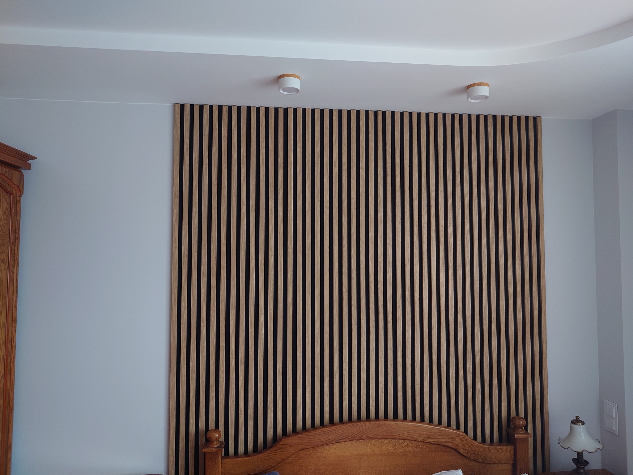 Ściana sypialni z drewnianymi lamelami w pionowym układzie, oświetlona dwoma okrągłymi lampami sufitowymi, widoczny fragment drewnianego wezgłowia łóżka i lampka nocna