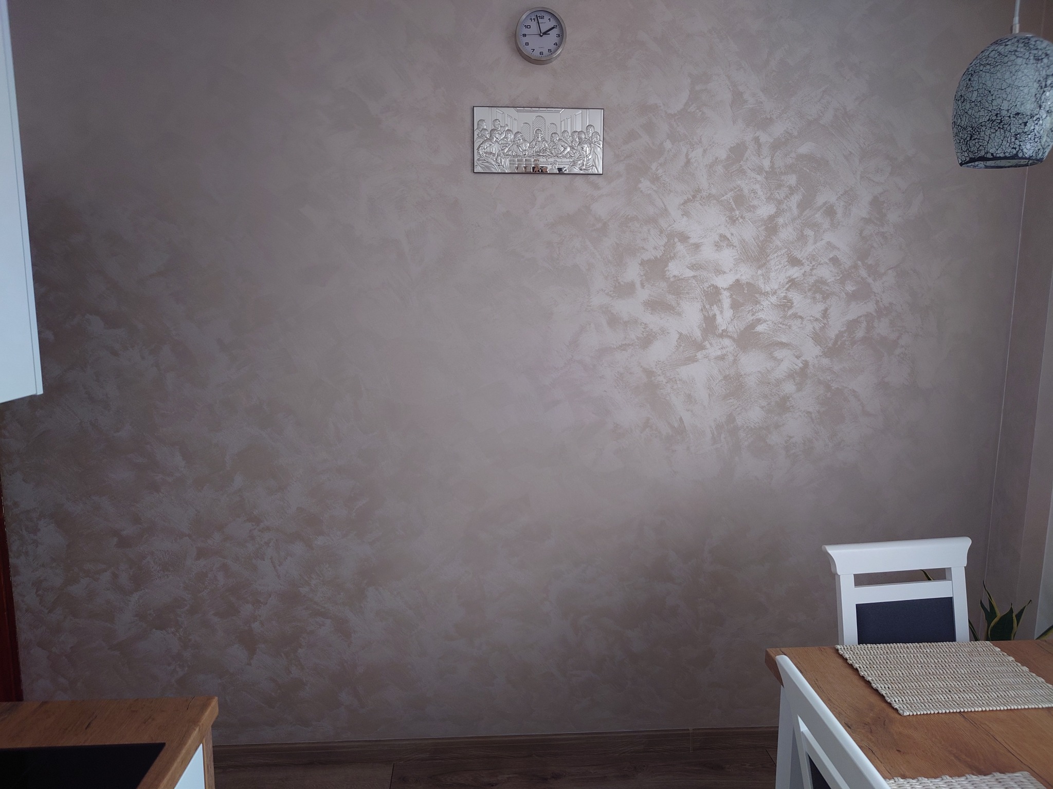 Ściana z dekoracyjnym tynkiem w odcieniach beżu i brązu, zegar i srebrny obrazek z Ostatnią Wieczerzą na ścianie, fragment stołu i krzesła w rogu pokoju, wisząca lampa.