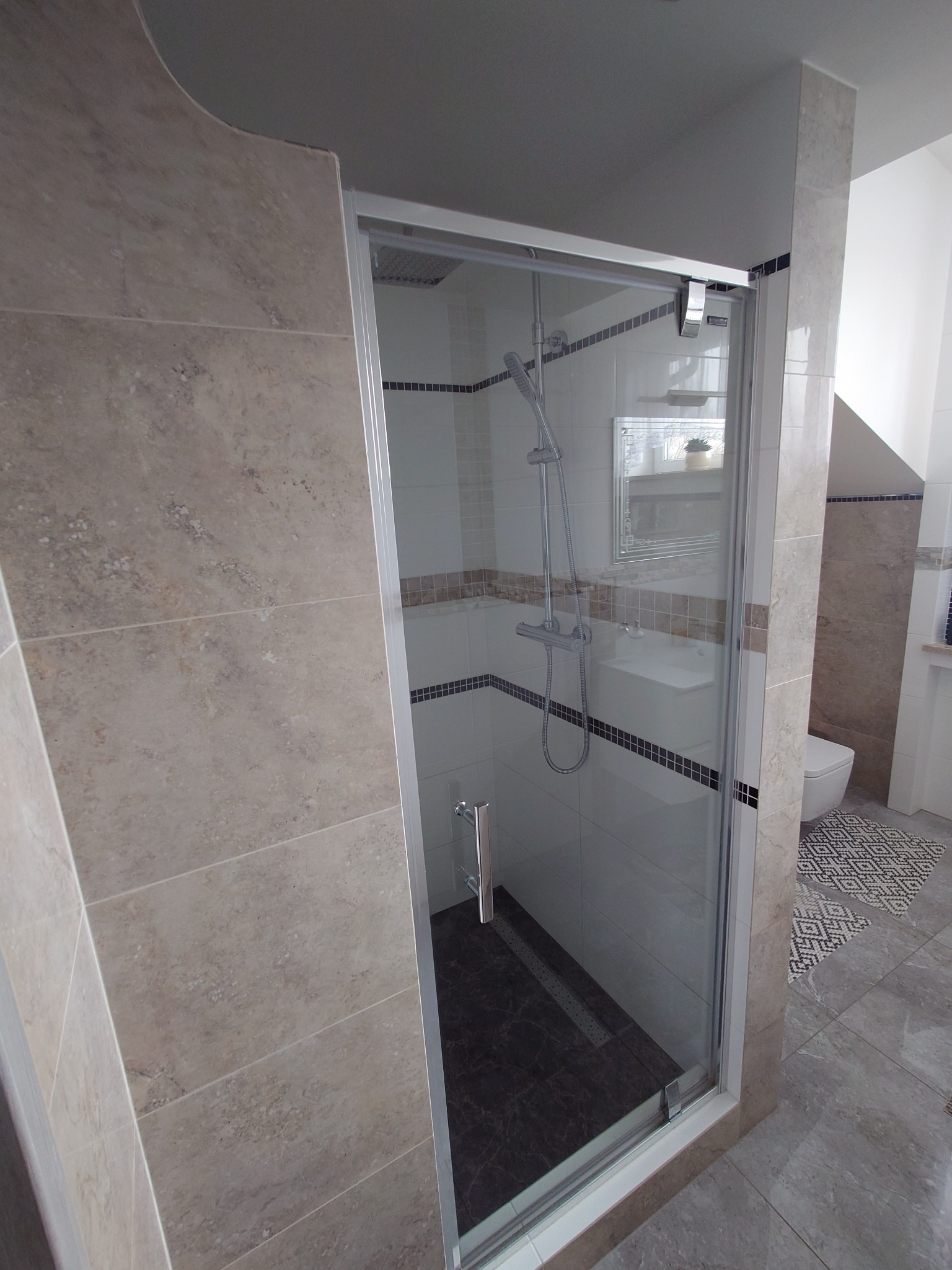 Nowoczesna kabina prysznicowa z przesuwnymi drzwiami i deszczownicą, wykończona płytkami w odcieniach beżu i szarości, z widocznym odpływem liniowym w podłodze.