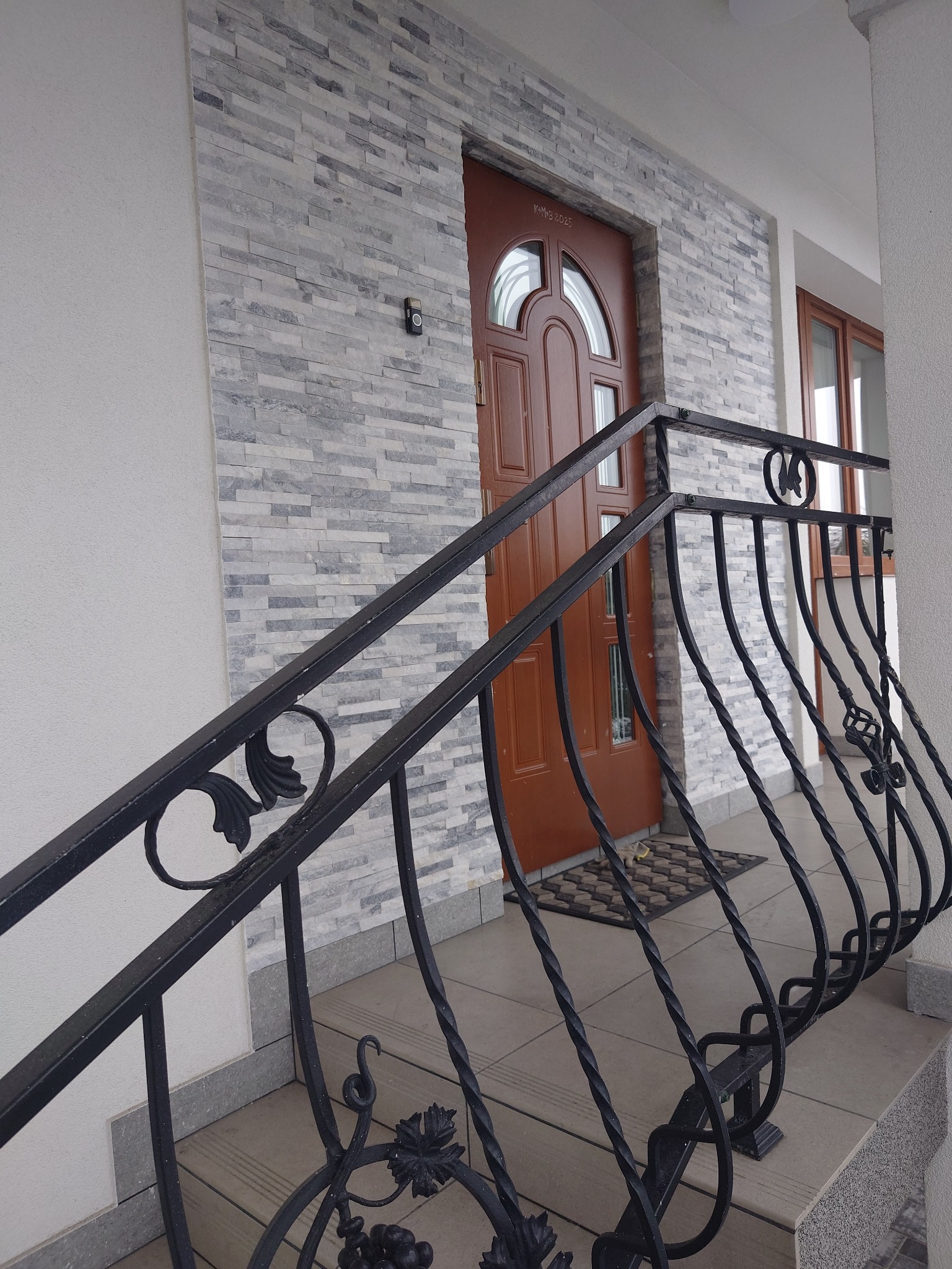 Wejście do domu z brązowymi drzwiami, wykończone szarym kamieniem elewacyjnym, ozdobione czarną, kutą balustradą z motywem winogron.