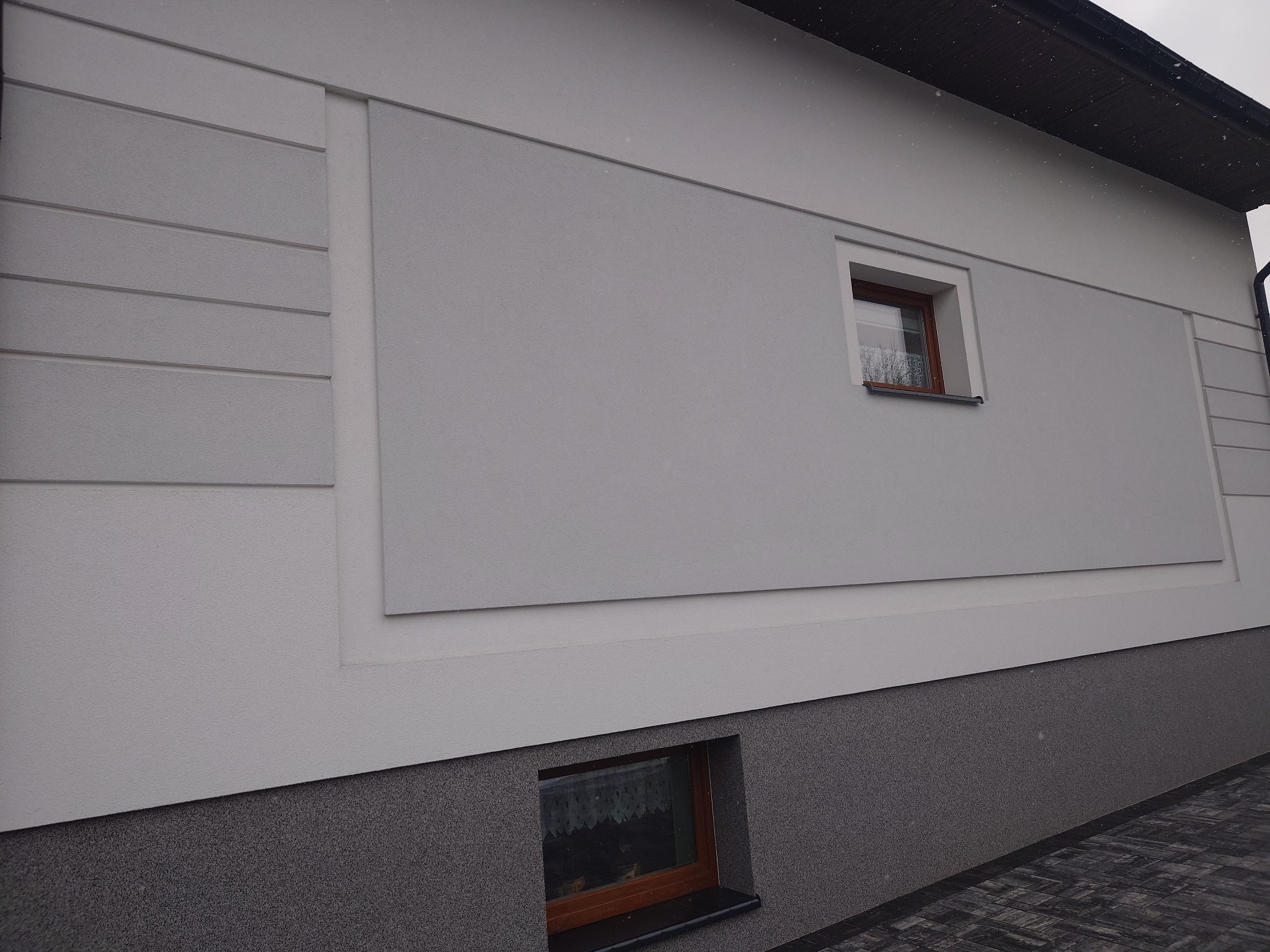 Fragment ocieplonej elewacji domu z dwoma oknami, z dekoracyjnymi poziomymi pasami i ciemniejszym cokołem.