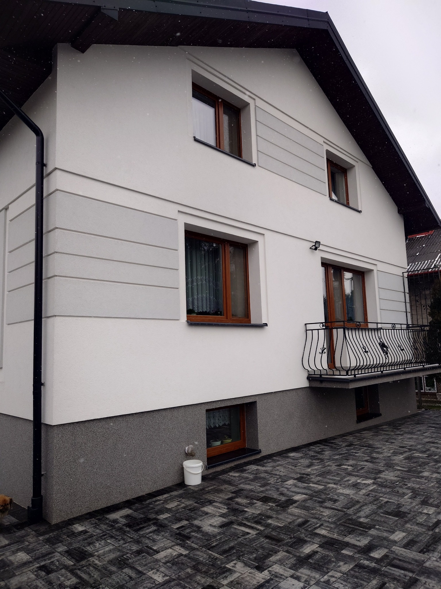 Dwupiętrowy dom jednorodzinny po świeżym ociepleniu elewacji w Radomiu, z widoczną strukturą tynku i dekoracyjnymi szarymi pasami, balkon z kutą balustradą, kostka brukowa na podjeździe i białe...