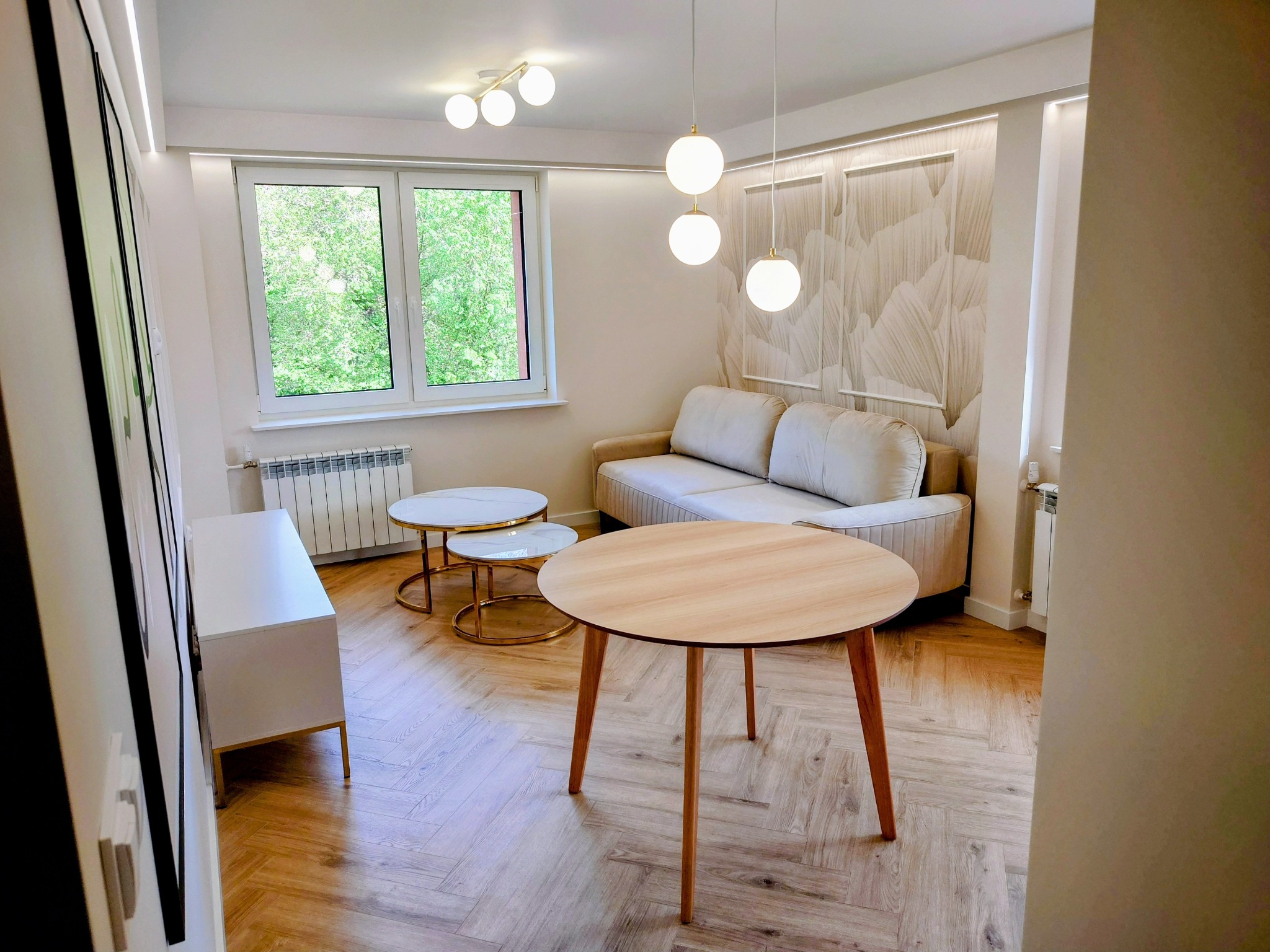 Jasne wnętrze po wykończeniu: parkiet w jodełkę, okrągły stół, sofa, stoliki kawowe, dekoracyjna ściana z motywem liści, nowoczesne lampy.