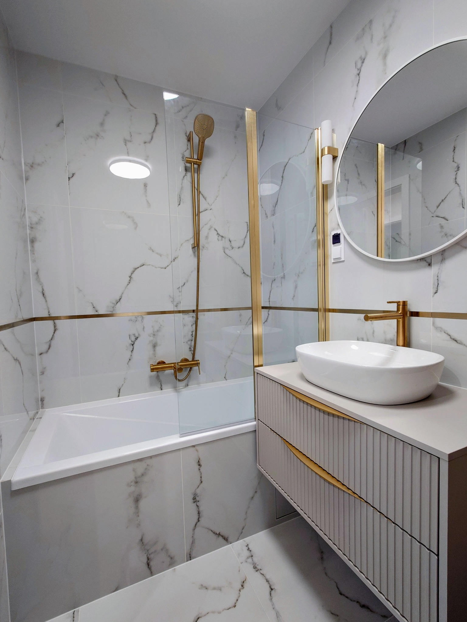 Nowoczesna łazienka z wanną i kabiną prysznicową oddzieloną szybą, wykończona białymi płytkami imitującymi marmur z delikatnymi złotymi akcentami.