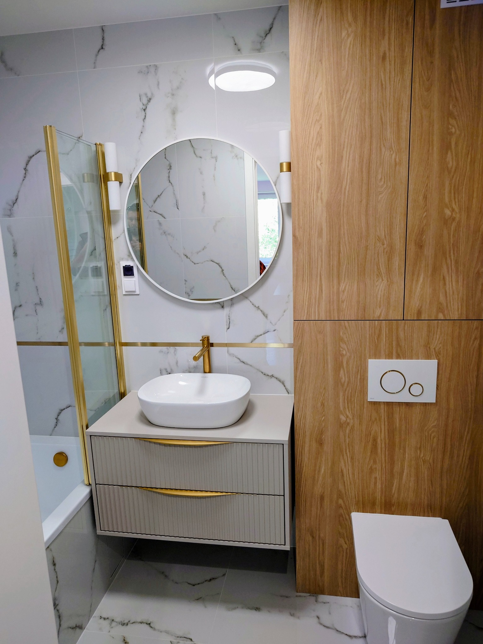 Wykończona łazienka w bieli i drewnie: umywalka na szafce z frezowanymi frontami, okrągłe lustro, kabina prysznicowa w złotych ramach, WC z drewnianą zabudową.