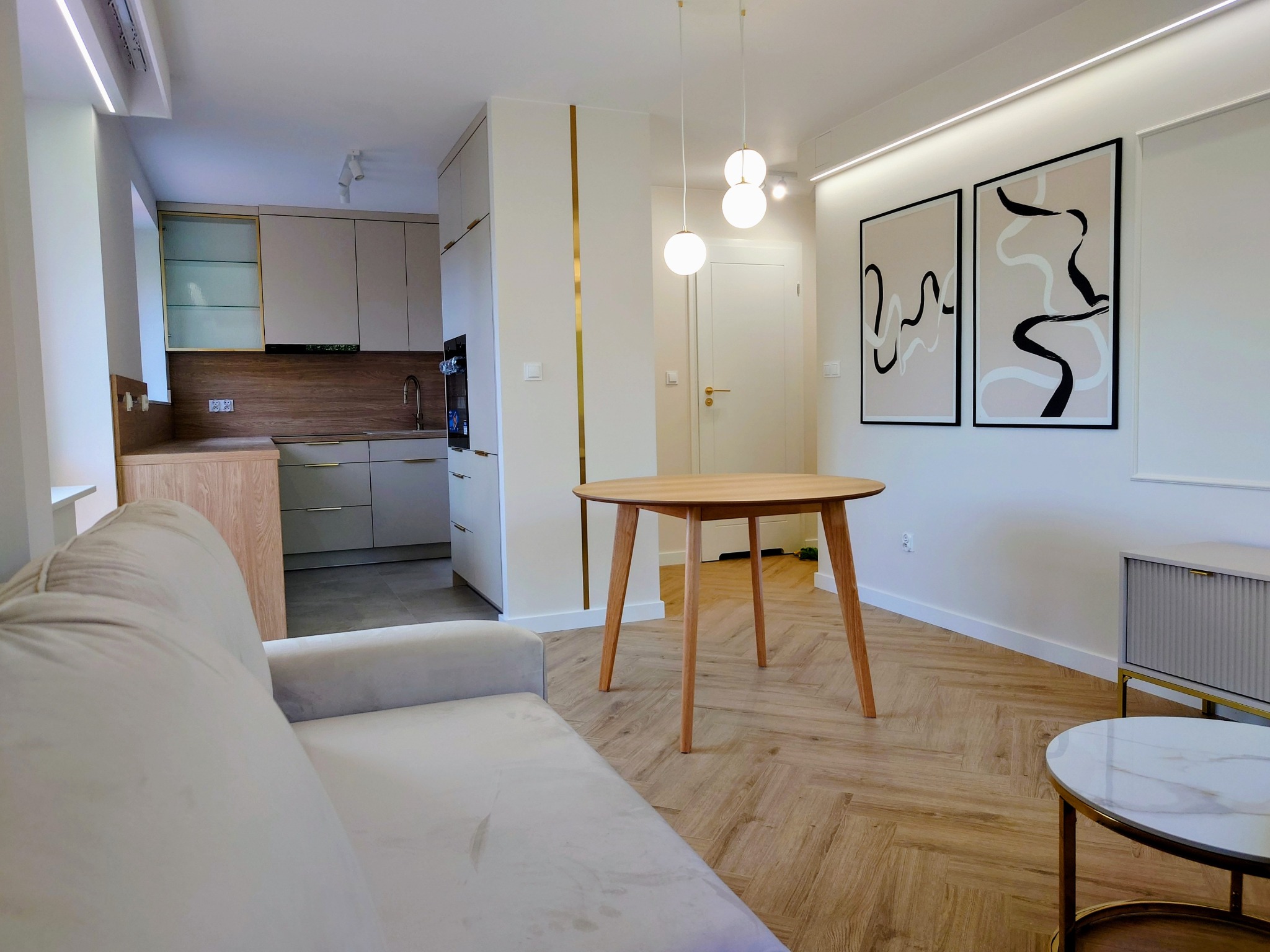 Jasne wnętrze po remoncie: parkiet w jodełkę, okrągły stół z drewna, beżowa sofa i minimalistyczne grafiki na ścianie. Kuchnia w tle, z zabudową w odcieniach beżu i drewna.