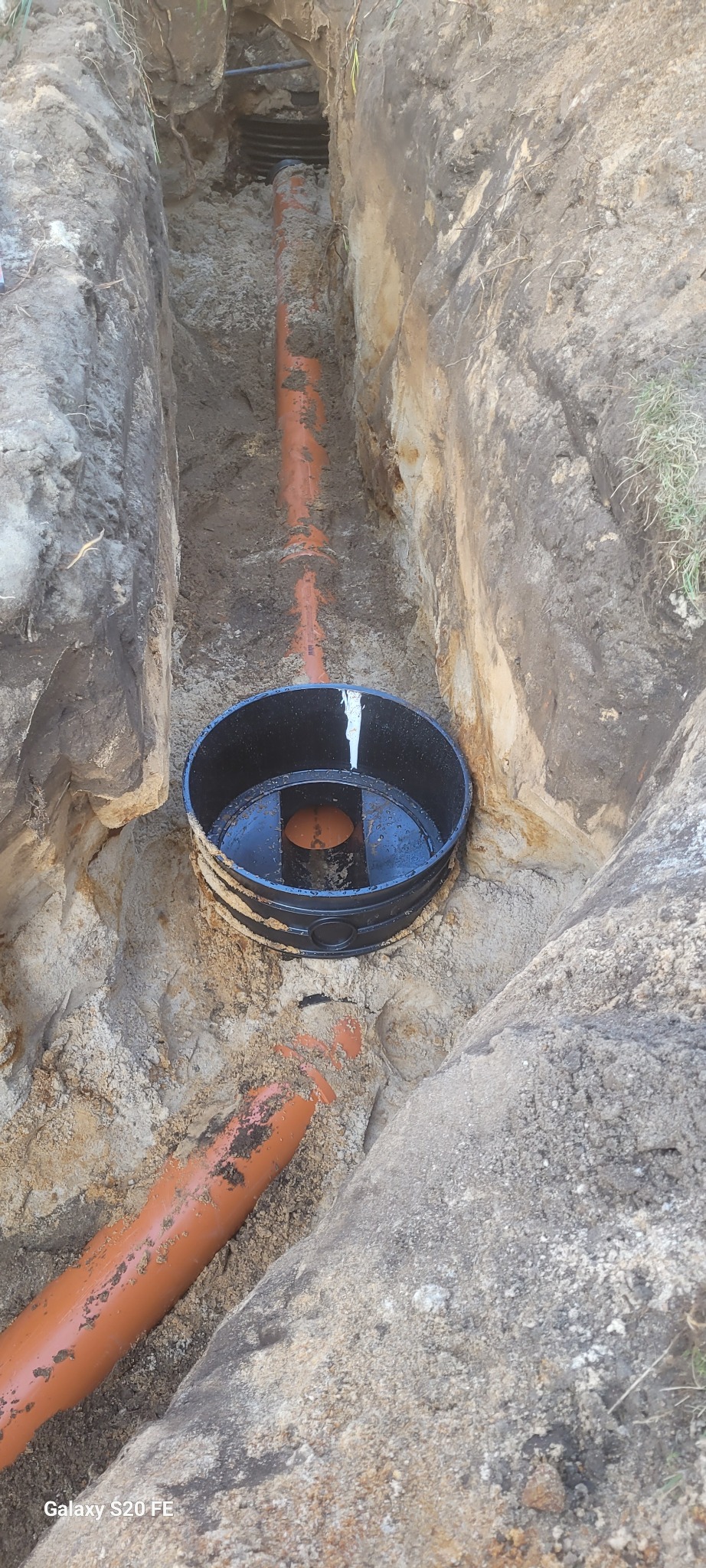 Wykop z pomarańczowymi rurami kanalizacyjnymi i czarną studzienką kanalizacyjną w trakcie montażu.