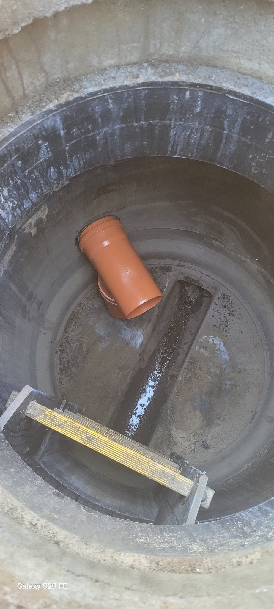 Wnętrze betonowej studni kanalizacyjnej z pomarańczową rurą PCV, drabinką i śladami wody na dnie.