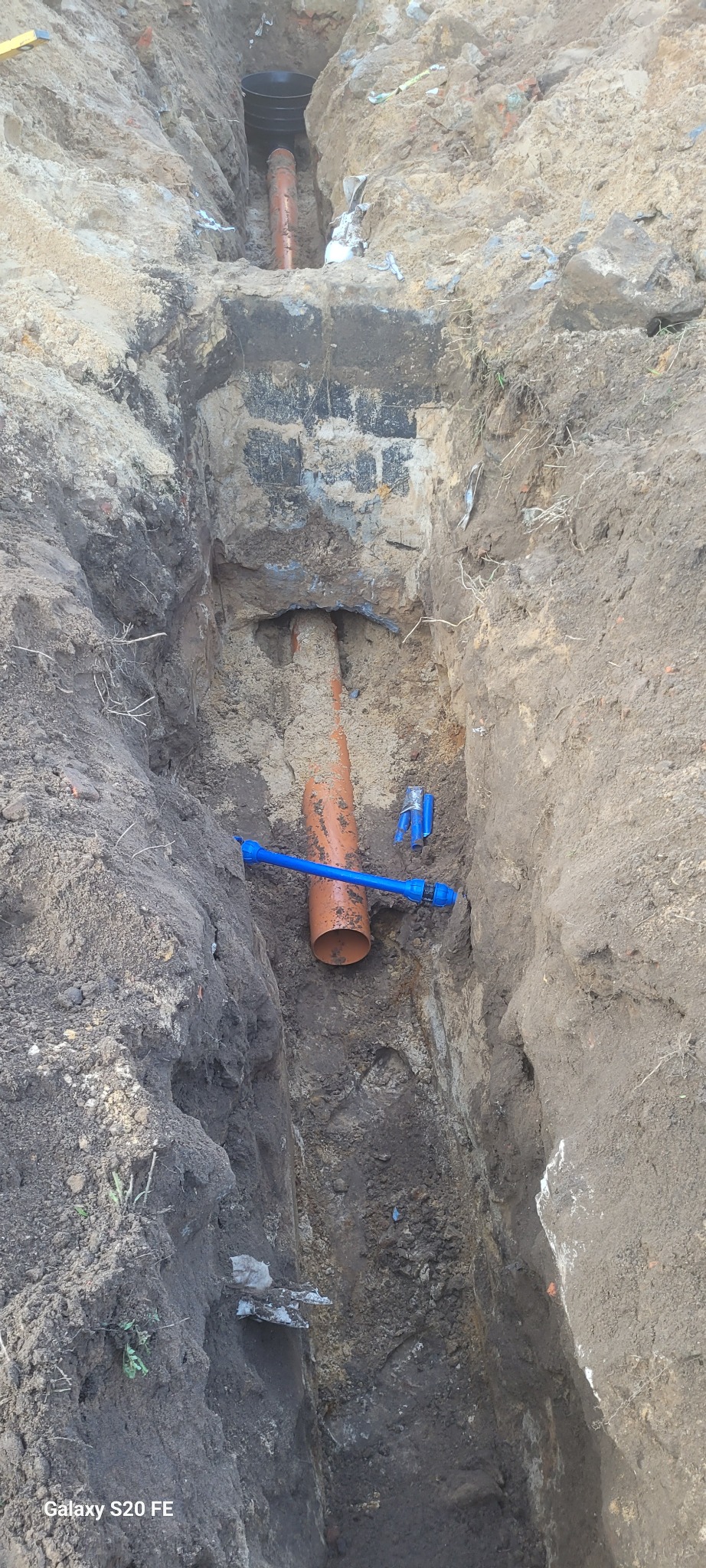 Wykop z pomarańczową rurą i niebieskimi złączkami w ziemi, widoczne warstwy gruntu i fragment starego muru z cegieł.