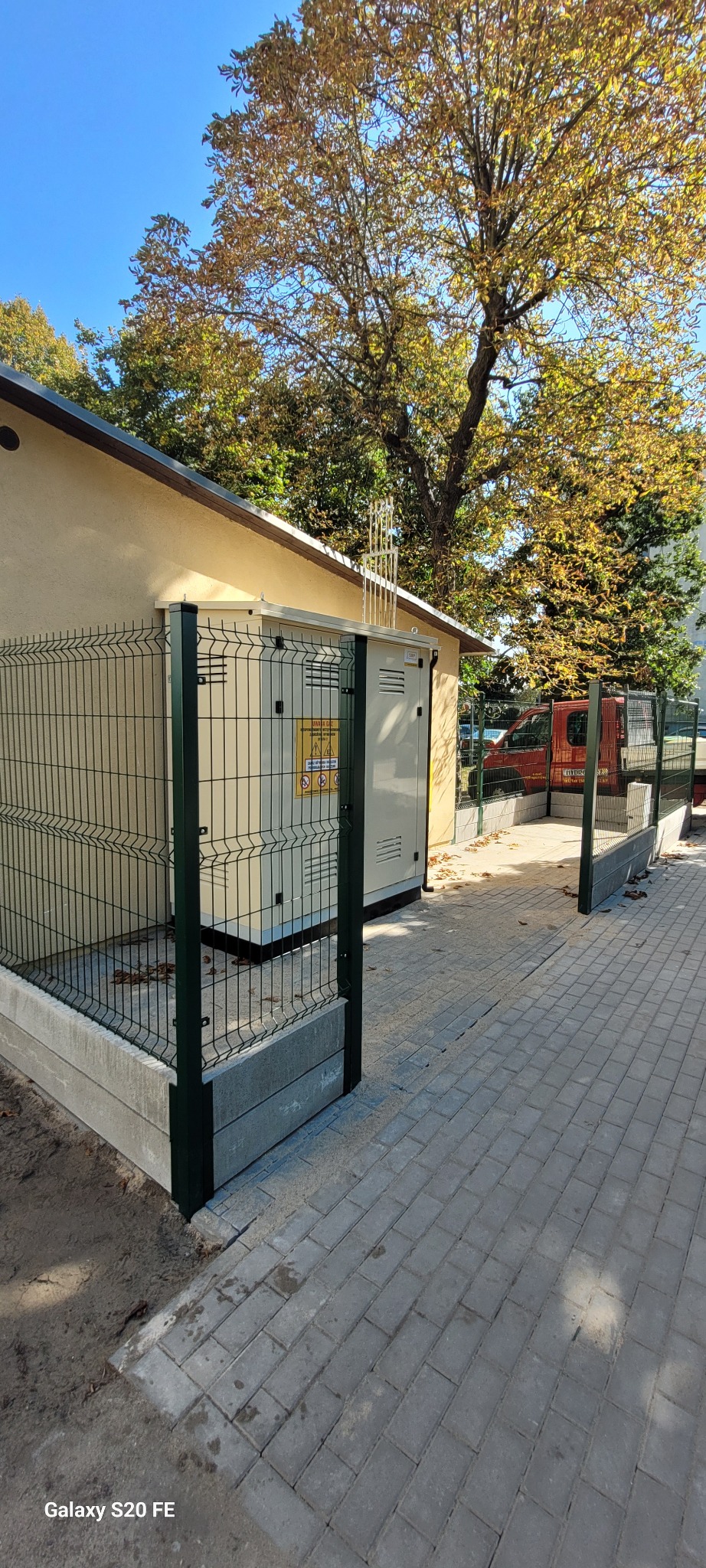 Zewnętrzna szafka gazowa zabezpieczona ogrodzeniem panelowym na betonowej podmurówce, widok z boku na tle budynku i drzew