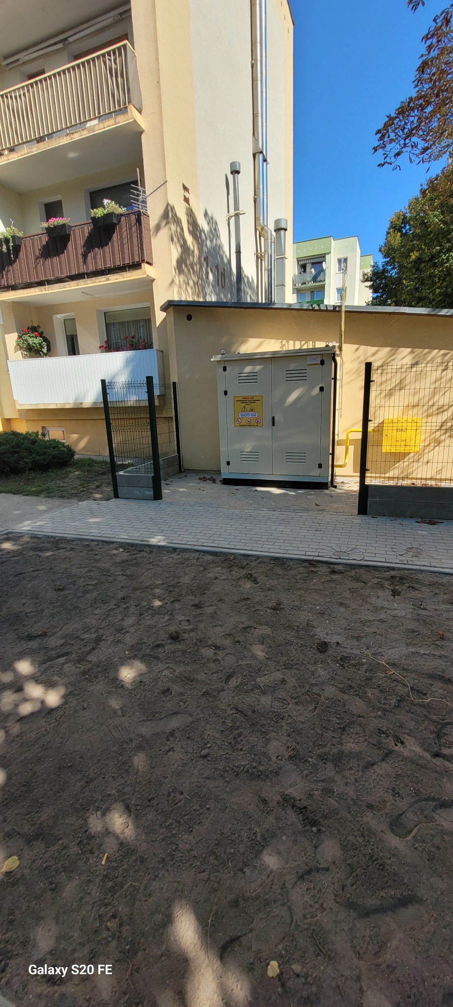 Zewnętrzna szafka gazowa z żółtymi oznaczeniami ostrzegawczymi, umieszczona przy budynku mieszkalnym z balkonami i metalowymi kominami, otoczona siatką i metalowym ogrodzeniem.