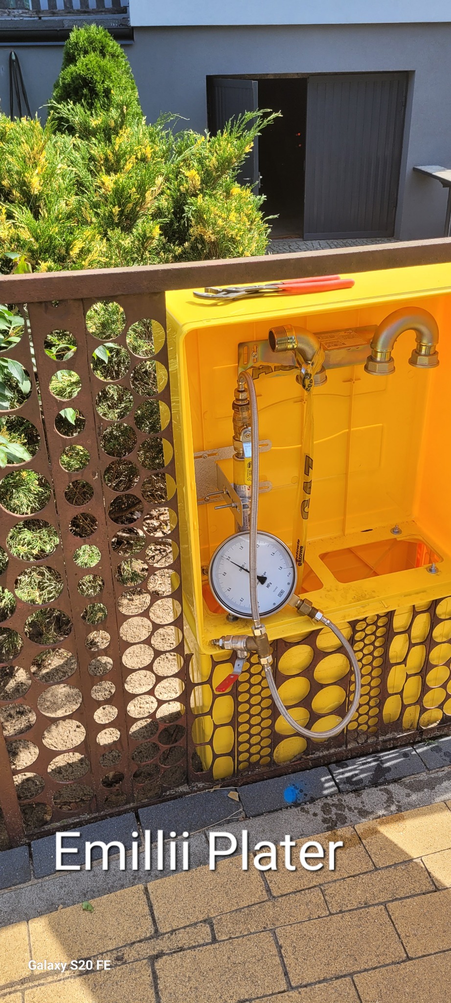 Żółta skrzynka z zamontowanymi przyłączami gazowymi i manometrem, widoczne narzędzia na górze, tło z zielenią i budynkiem.