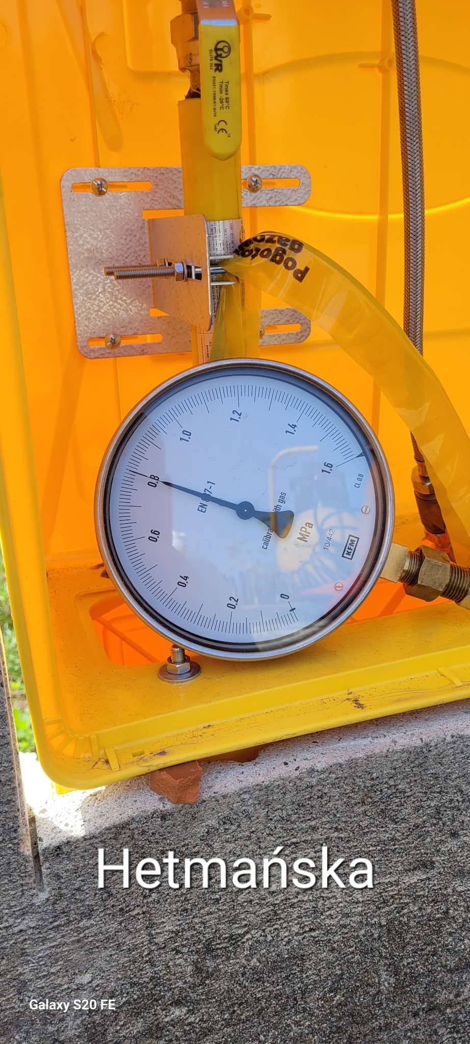 Manometr w żółtej skrzynce pomiarowej, wskazujący ciśnienie robocze, z widocznymi elementami instalacji gazowej, takimi jak zawór i elastyczny przewód.