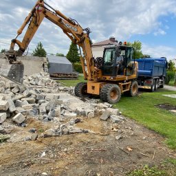 KOP-TRANS DAWID SIWIEC - Żółta koparka CASE z operatorem, gruz po rozbiórce budynku i wywrotka z niebieską skrzynią na tle zabudowań i zieleni. Rozbiórka w toku, widoczne elementy konstrukcyjne.