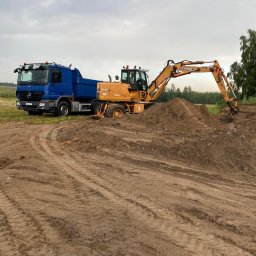 KOP-TRANS DAWID SIWIEC - Niebieska ciężarówka i żółta koparka CASE WX120 na placu budowy, w tle zielone drzewa i pochmurne niebo. Widoczne ślady opon na ziemi.