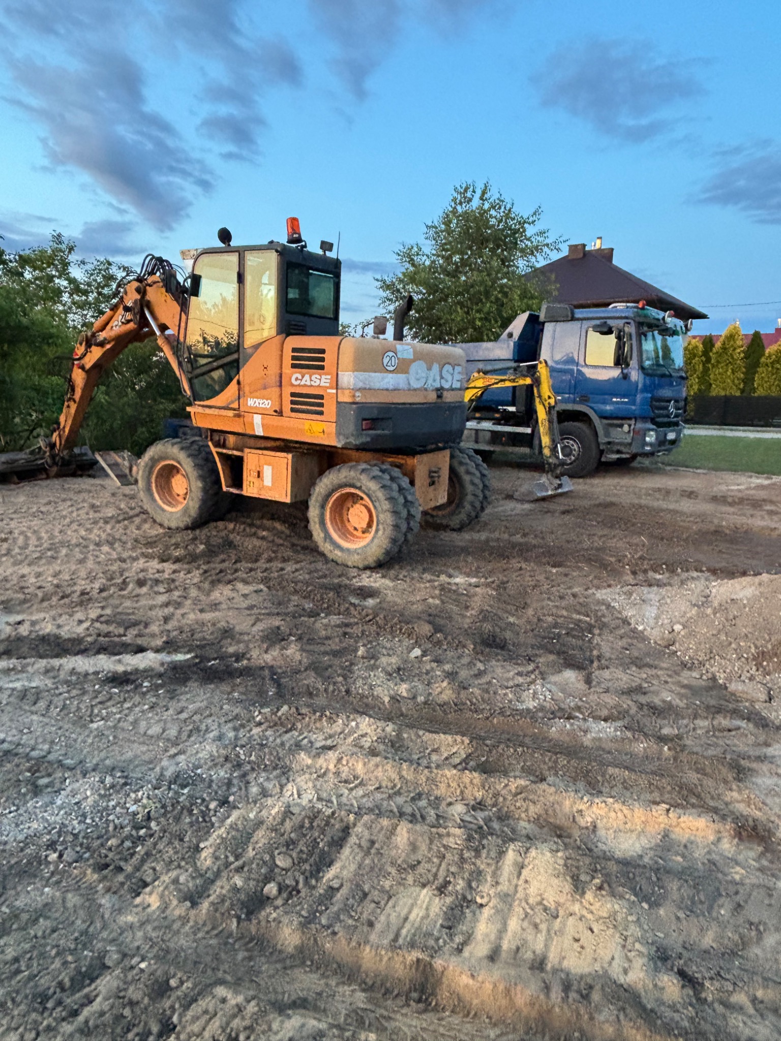 Pomarańczowa koparka CASE WX120 i niebieska ciężarówka na placu budowy z ubitym gruntem, drzewa i dom w tle, pochmurne niebo.