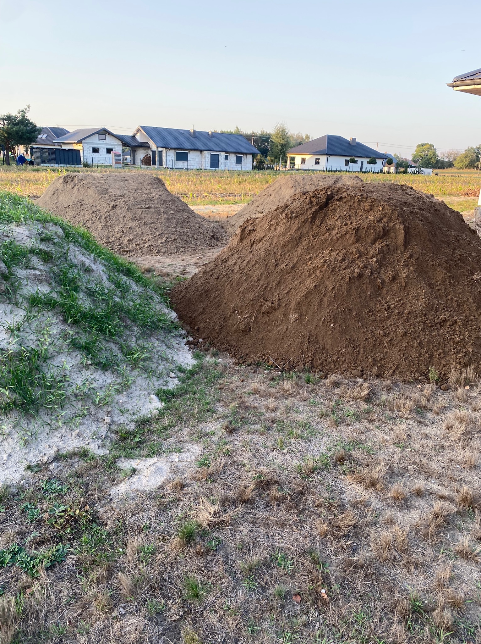 Trzy pryzmy ziemi o różnej barwie (szara, brązowa, ciemnobrązowa) na tle budynków w trakcie budowy. Ziemia częściowo porośnięta trawą.