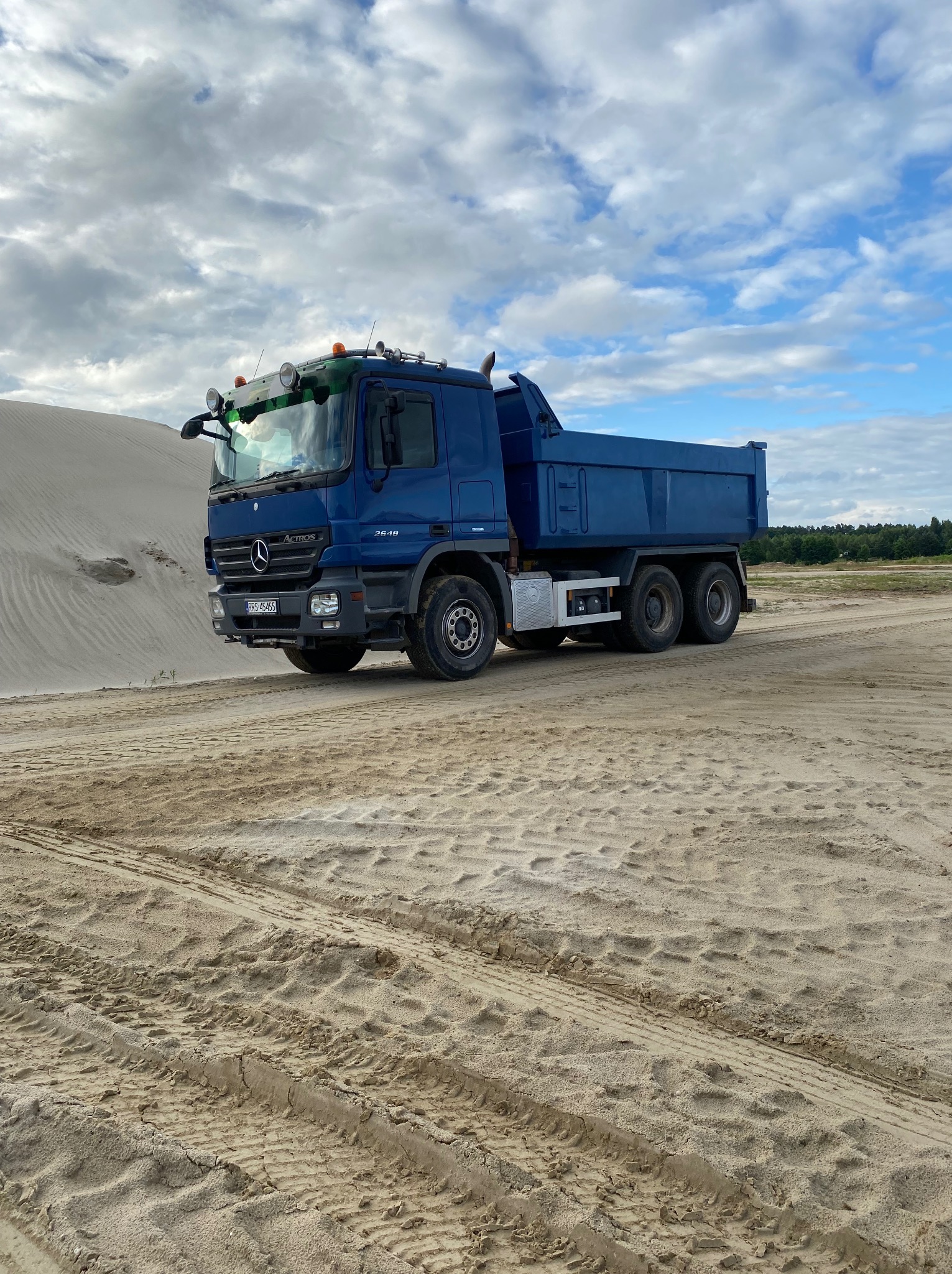 Niebieska wywrotka Mercedes Actros na piaszczystym terenie z widocznymi śladami opon i wydmą w tle, pod błękitnym niebem z chmurami.