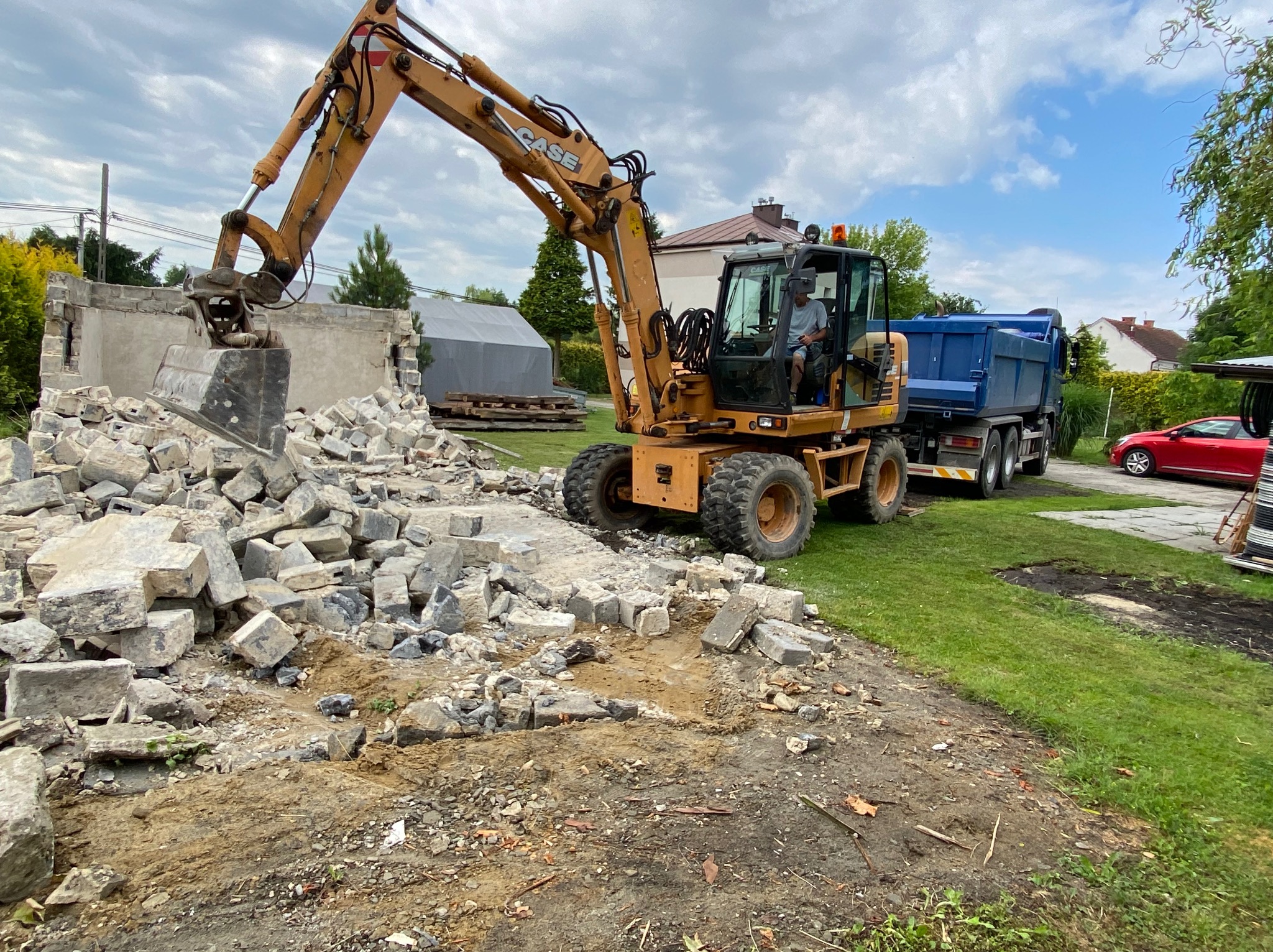 Żółta koparka CASE z operatorem, gruz po rozbiórce budynku i wywrotka z niebieską skrzynią na tle zabudowań i zieleni. Rozbiórka w toku, widoczne elementy konstrukcyjne.