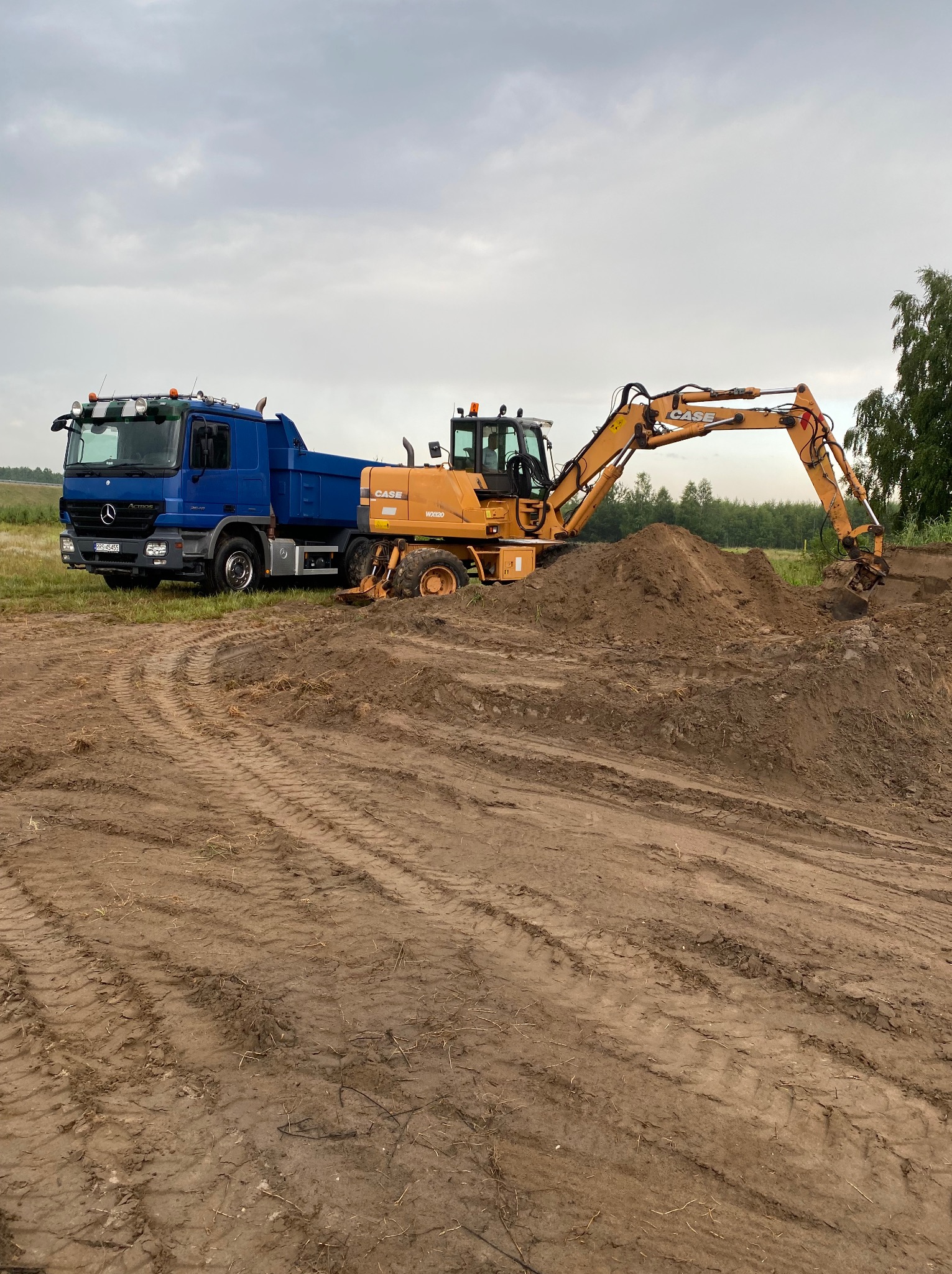 Niebieska ciężarówka i żółta koparka CASE WX120 na placu budowy, w tle zielone drzewa i pochmurne niebo. Widoczne ślady opon na ziemi.