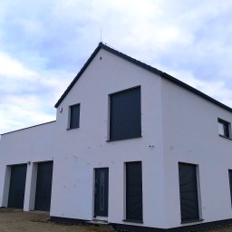 Wieczorek Budownictwo - Nowoczesny, biały dom z szarymi roletami i garażem, widok z dołu. Na dachu widoczny piorunochron. Świeżo wykończona elewacja, czyste linie architektoniczne.