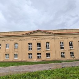 Wieczorek Budownictwo - Elewacja budynku w klasycystycznym stylu, beżowa fasada z rzędami okien i dekoracyjnymi elementami architektonicznymi, widok z perspektywy trawnika.