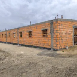 Budowa domu z czerwonej cegły: ściany wzniesione, otwory okienne przygotowane, widoczne zbrojenia. Tło: pochmurne niebo, teren budowy.