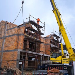 Wieczorek Budownictwo - Budowa domu z cegły ceramicznej: murarz w pomarańczowym kombinezonie na rusztowaniu, dźwig budowlany w tle. Widoczne materiały budowlane i ogrodzenie placu budowy.