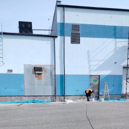 Wieczorek Budownictwo - Pracownik maluje elewację budynku na niebiesko, widoczne rusztowania i drabina. Część ściany już pomalowana, reszta przygotowana pod malowanie.