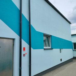 Wieczorek Budownictwo - Pomalowana na dwa odcienie błękitu ściana budynku z metalowymi rurami pionowymi, oknem i metalowymi drzwiami. Widoczne znaki ostrzegawcze.