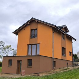 Wieczorek Budownictwo - Dwukondygnacyjny dom z pomarańczową elewacją i brązowym cokołem, widoczny z perspektywy trawnika. W tle drzewa i sąsiedni budynek. Pochmurne niebo.