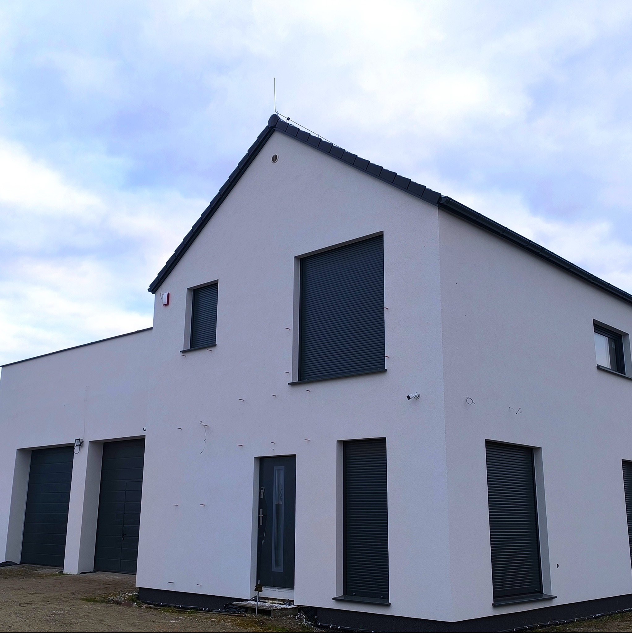 Nowoczesny, biały dom z szarymi roletami i garażem, widok z dołu. Na dachu widoczny piorunochron. Świeżo wykończona elewacja, czyste linie architektoniczne.
