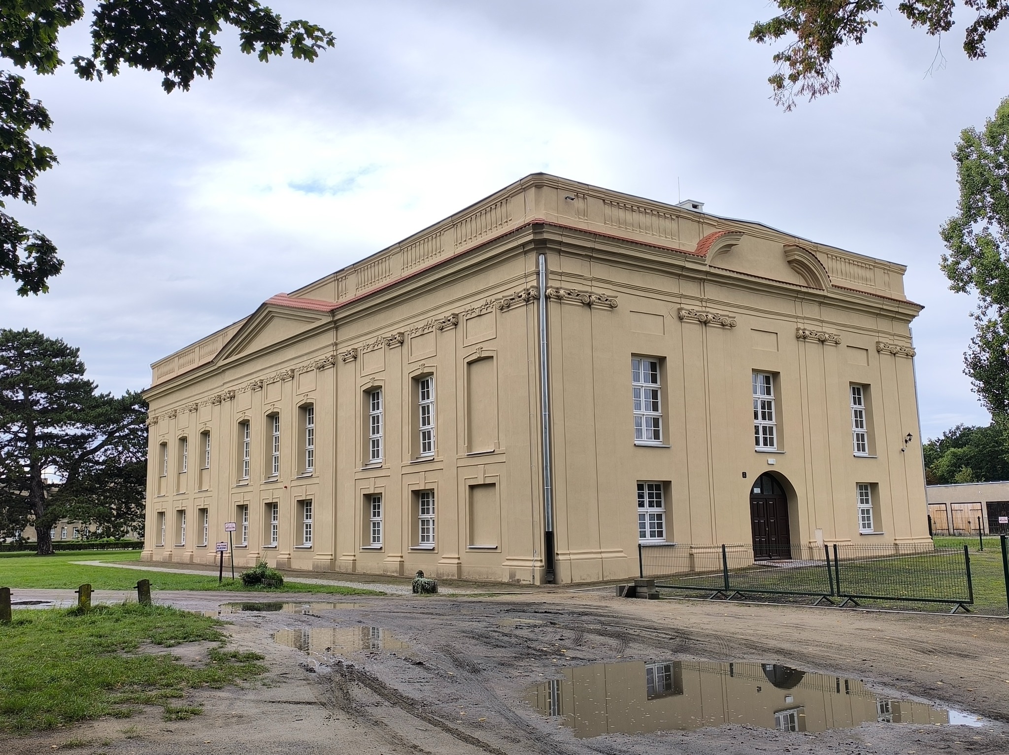 Elewacja odnowionego, beżowego budynku z białymi oknami, widoczna z zewnątrz. Na pierwszym planie kałuże odbijające budynek i pochmurne niebo. Ujęcie z dołu.