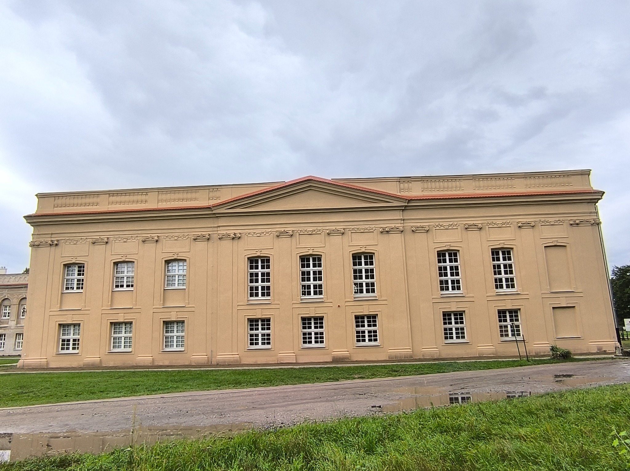 Elewacja budynku w klasycystycznym stylu, beżowa fasada z rzędami okien i dekoracyjnymi elementami architektonicznymi, widok z perspektywy trawnika.