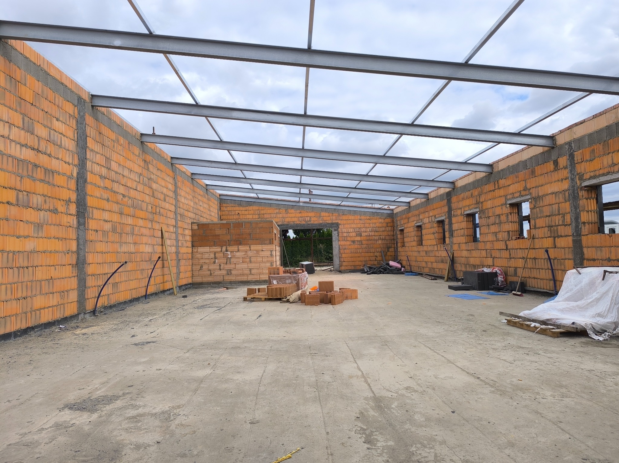 Wnętrze budynku w trakcie budowy: ściany z pomarańczowych bloczków, stalowa konstrukcja dachu, betonowa podłoga. Widoczne materiały budowlane i narzędzia.