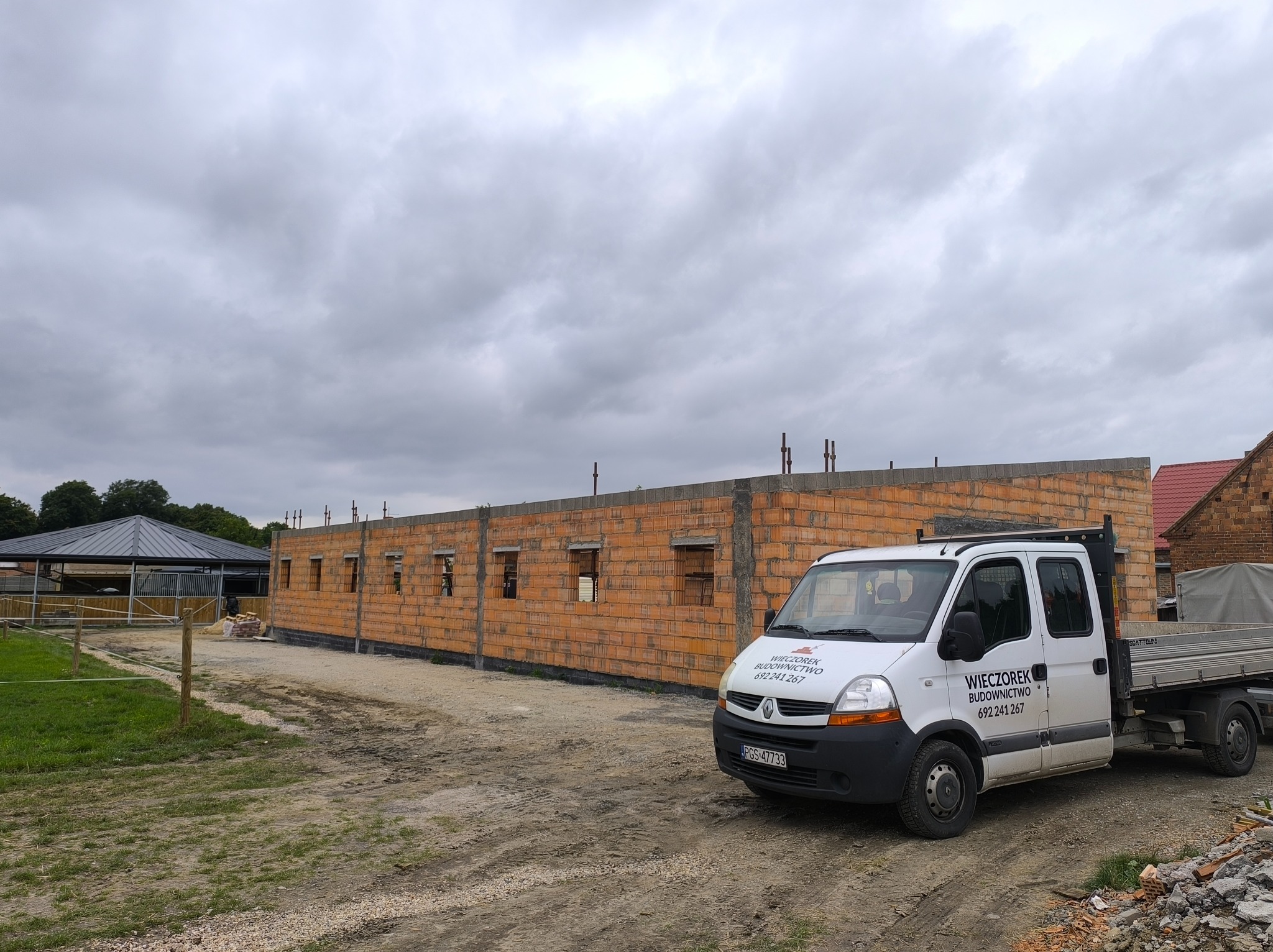 Budowa budynku z cegły ceramicznej, widoczny mur z otworami okiennymi, na pierwszym planie samochód dostawczy z logo firmy budowlanej. Pochmurne niebo.